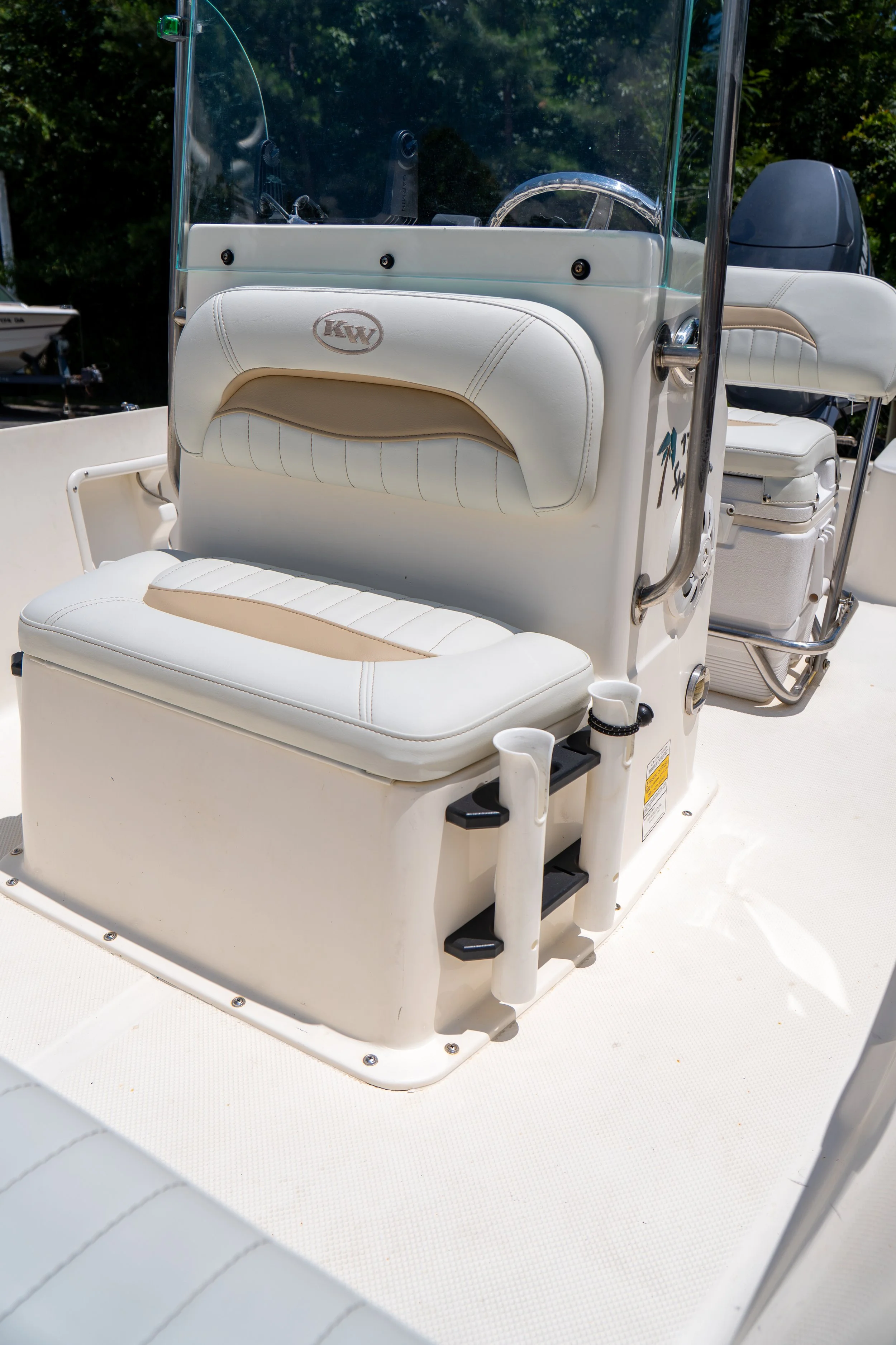 Front view of a white boat with beige seat padding, featuring a windshield, steering wheel, and a small mounted outboard motor at the rear. The boat appears to be docked or out on water under a clear sky.
