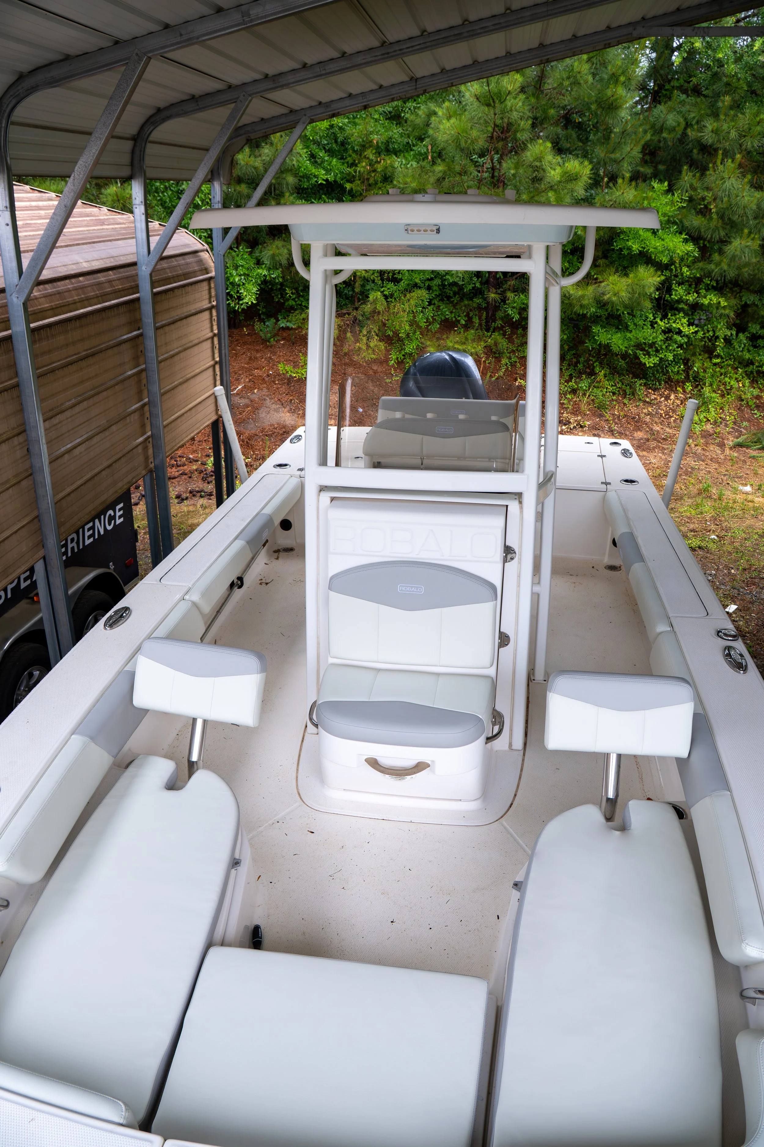 Top view of a white boat with several seats and a steering console, parked under a metal roof near green trees and bushes.