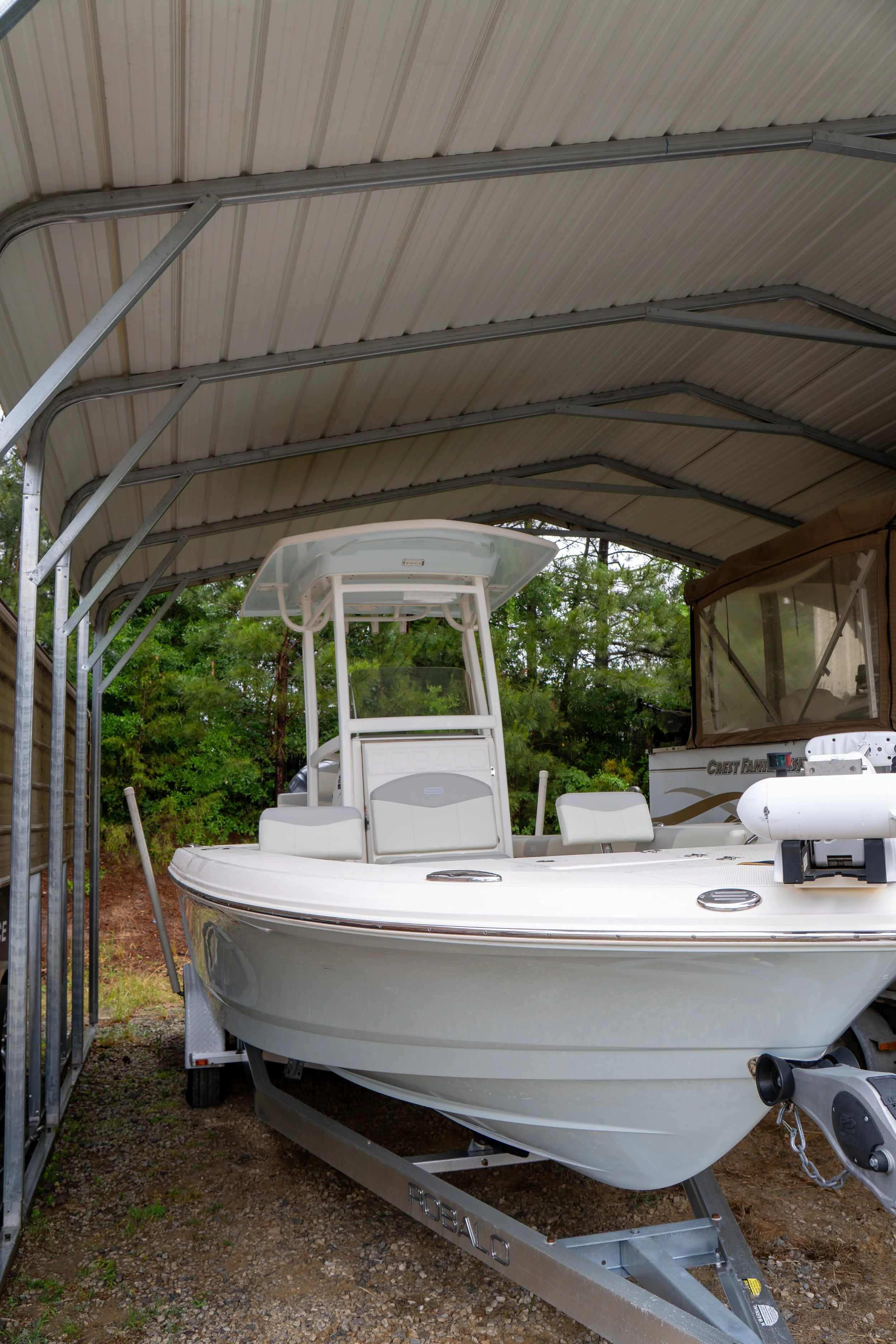 A white boat on a trailer parked under a metal-roof shelter with trees in the background.
