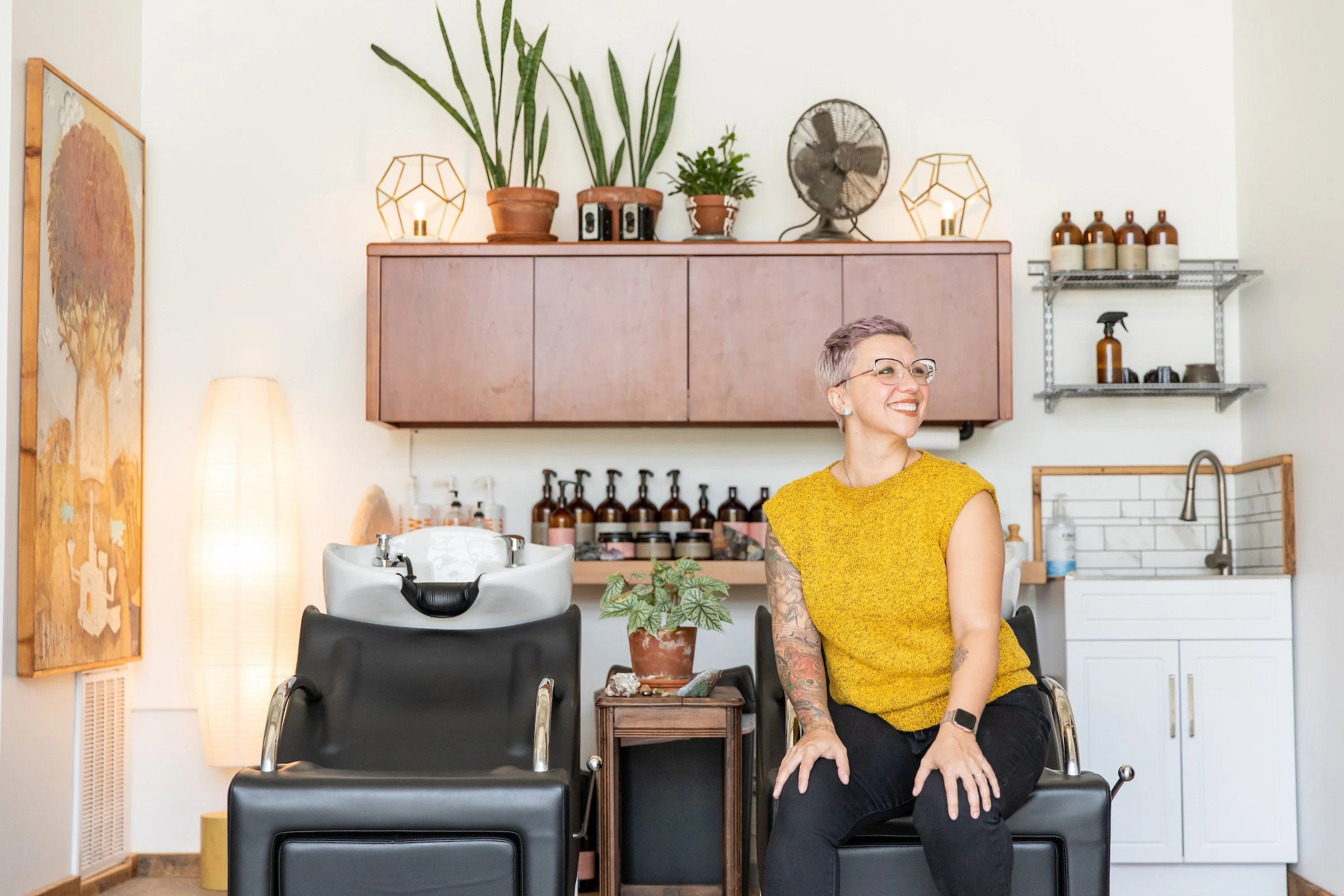 Woman with short purple hair smiling off camera and sitting in a shampoo chair in a salon.