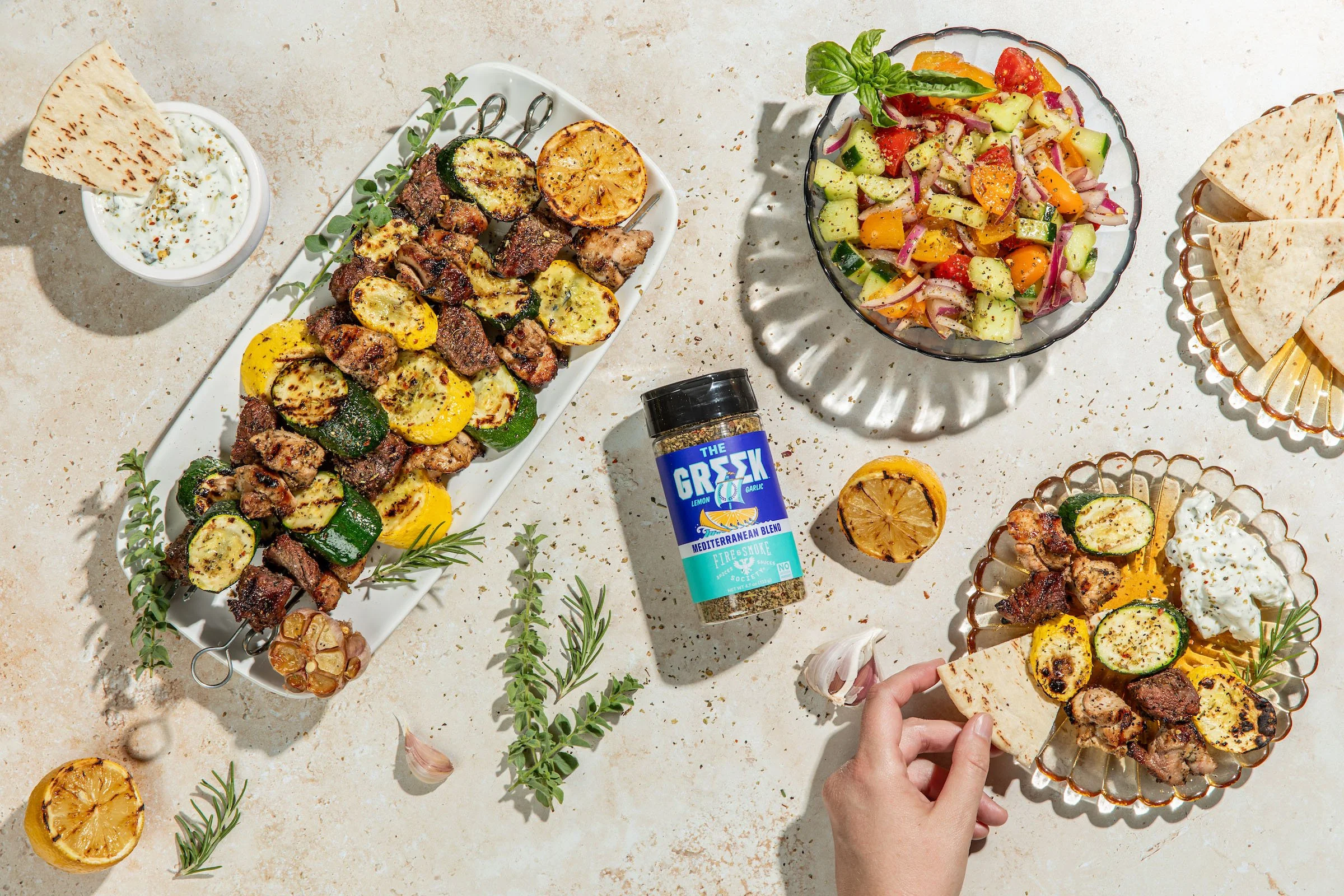 Table scene from above of a Greek seasoning bottle surrounded by platters and plates of food.