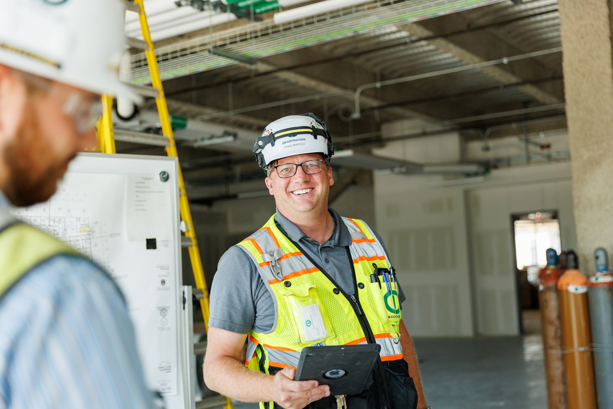 Construction worker with glasses and a safety helmet smiling, holding a tablet, at an indoor construction site with pipes and equipment.