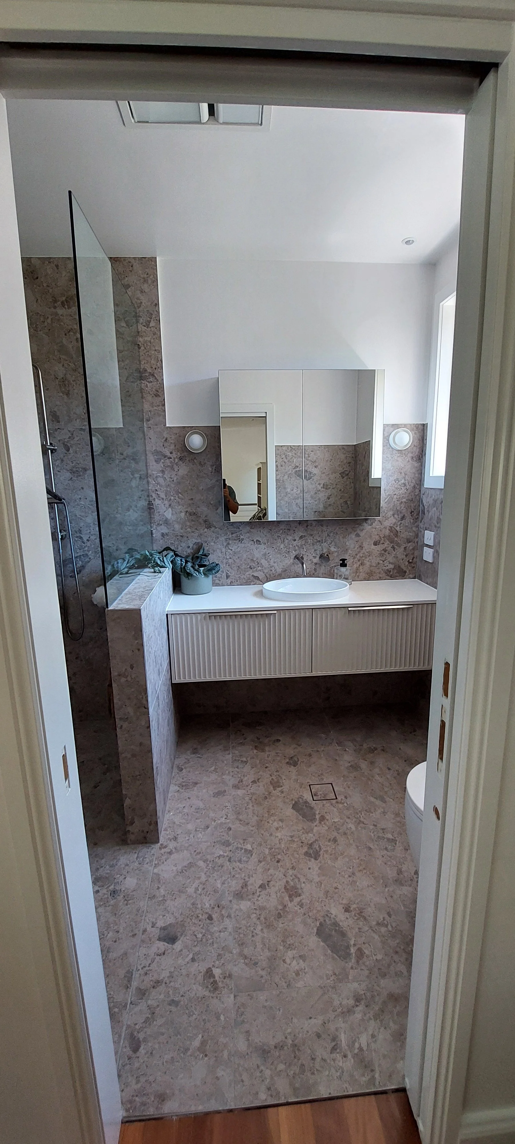 Modern bathroom with marble tiles, a floating vanity with a white sink, a large mirrored cabinet, a plant on the counter, and a shower area separated by a glass partition. There is a window on the right side, and the floor is also marble.
