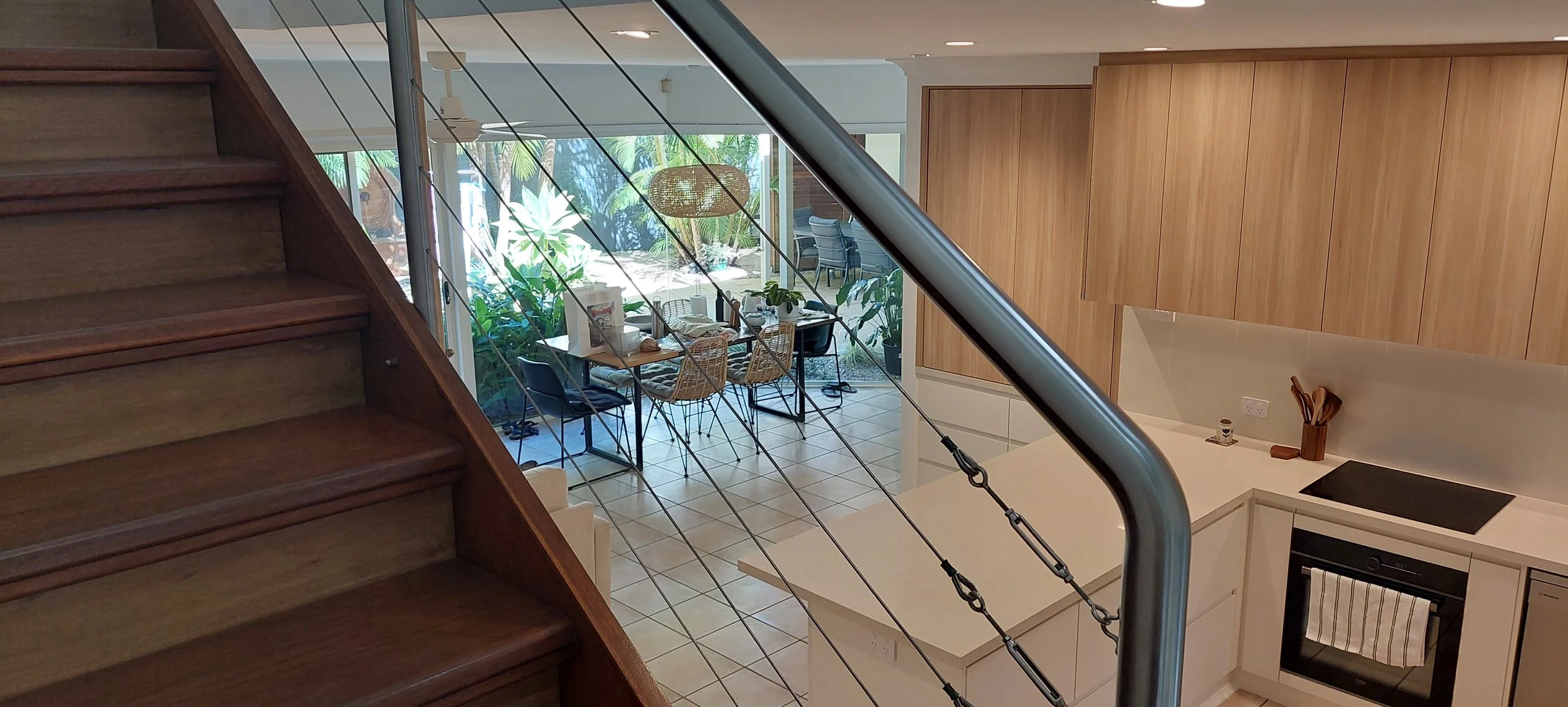 Interior view of a modern kitchen and dining area seen from the staircase, with wooden cabinets, a white countertop, and a table with chairs in front of a large window with greenery outside.