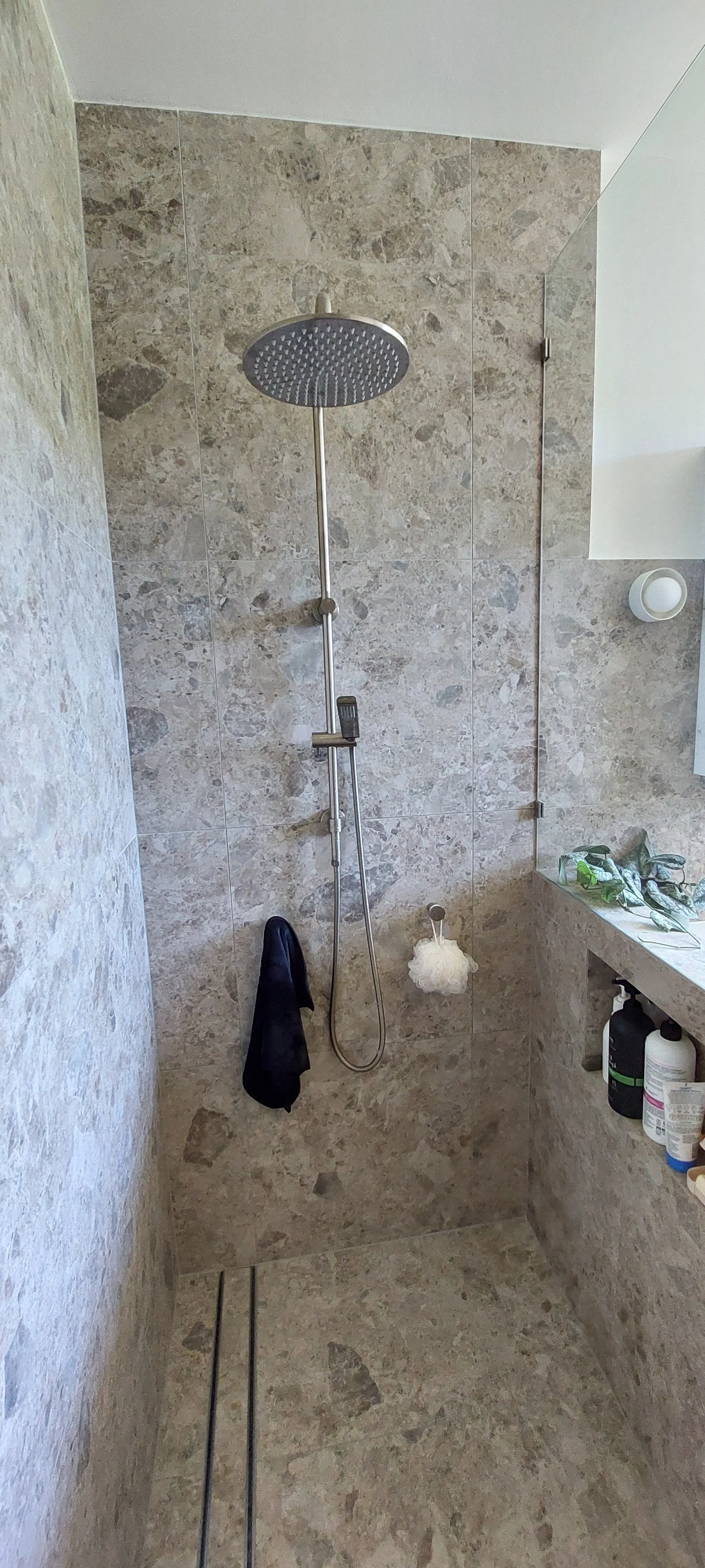 A modern shower with a large rainfall showerhead, a handheld shower wand, beige and gray marble tiles, a black towel hanging, a loofah, and a partially visible countertop with toiletries inside a bathroom.