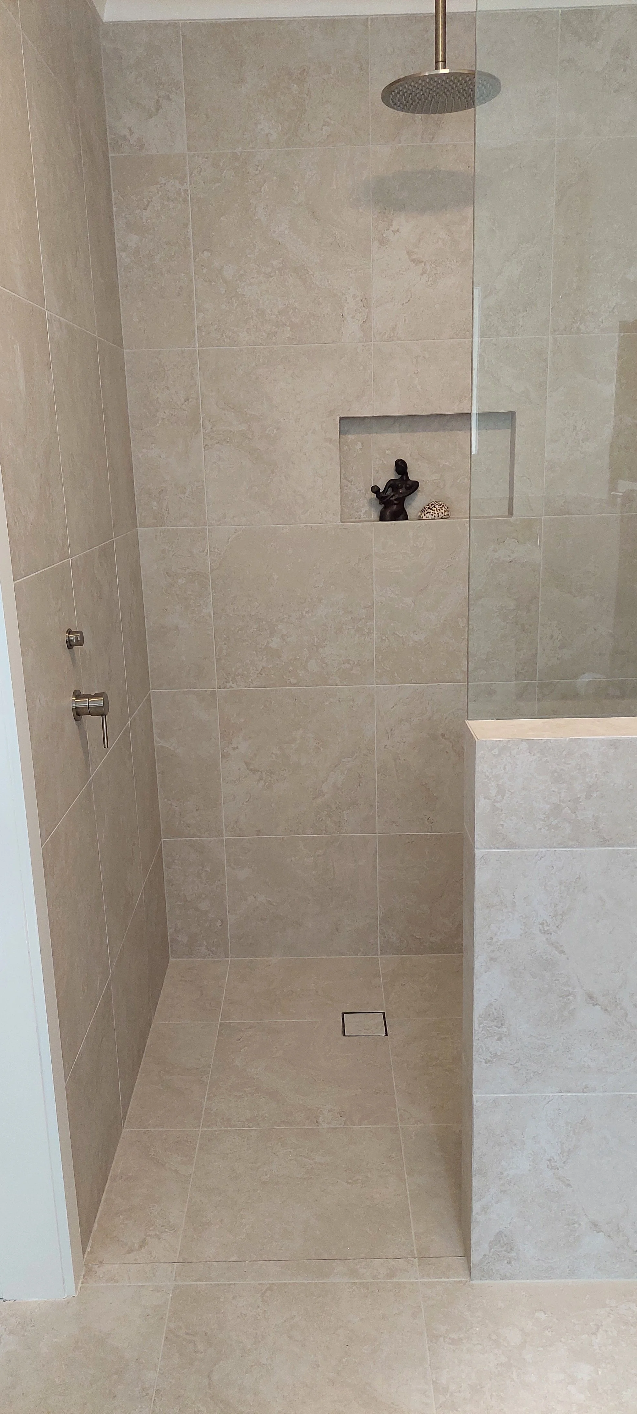 A modern walk-in shower with beige tiled walls and floor, a rain showerhead, a built-in niche with decorative items, and a partial glass partition.