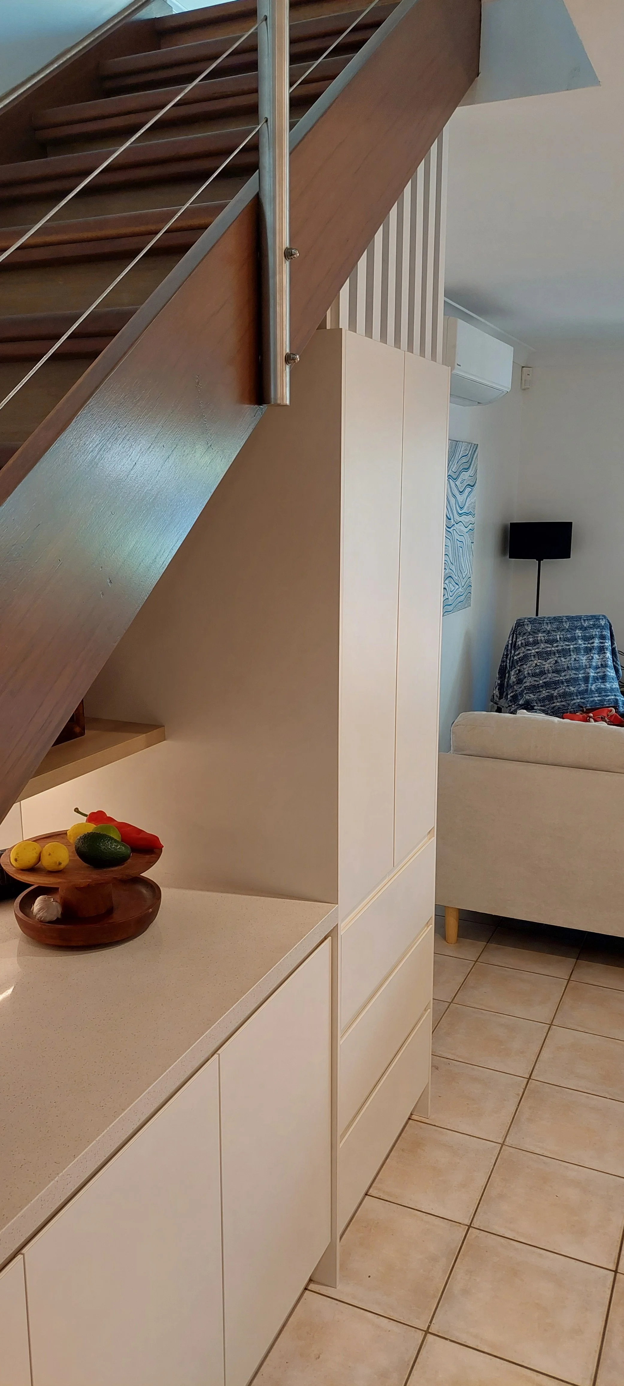 Wooden staircase with metal railing next to a white cabinet in a living space. A bowl of fruits, including lemons, an avocado, and a chili pepper, sits on the countertop. In the background, a beige sofa with a dark blue patterned blanket or cloth dra