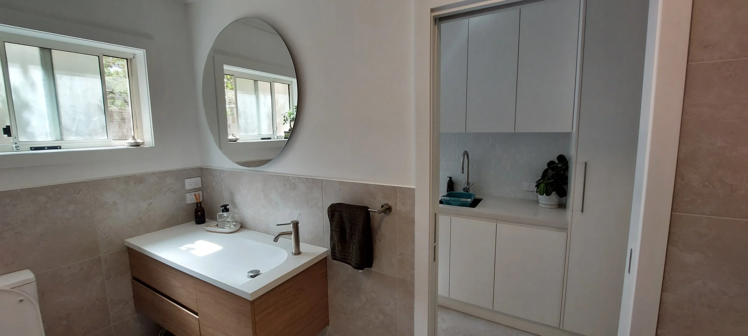 A modern bathroom with a white sink vanity, an oval mirror, a window with frosted glass, a small cabinet with a plant, and neutral-toned tiles.