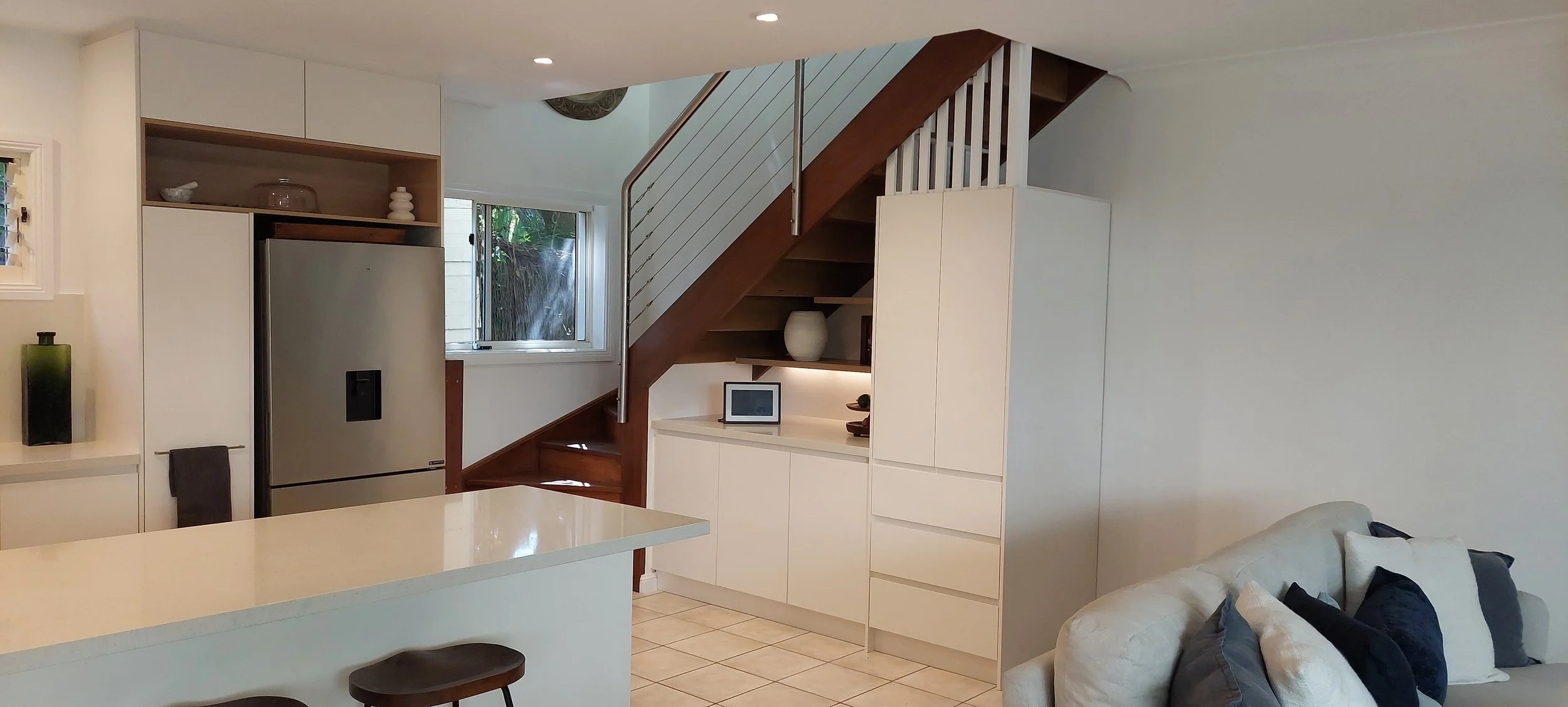Interior view of a modern home with a kitchen, staircase, and living area. Kitchen features white cabinetry, a stainless steel refrigerator, and a white island with two stools. A staircase with wooden steps and a metal railing leads upstairs. There i