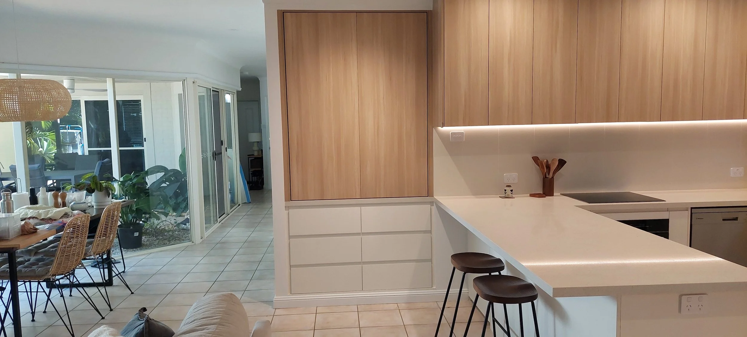 Open-plan kitchen and dining area with large wooden cabinets, white countertops, and a breakfast bar with three barstools. Adjacent to a glass sliding door, there are plants and a dining table with woven chairs inside.