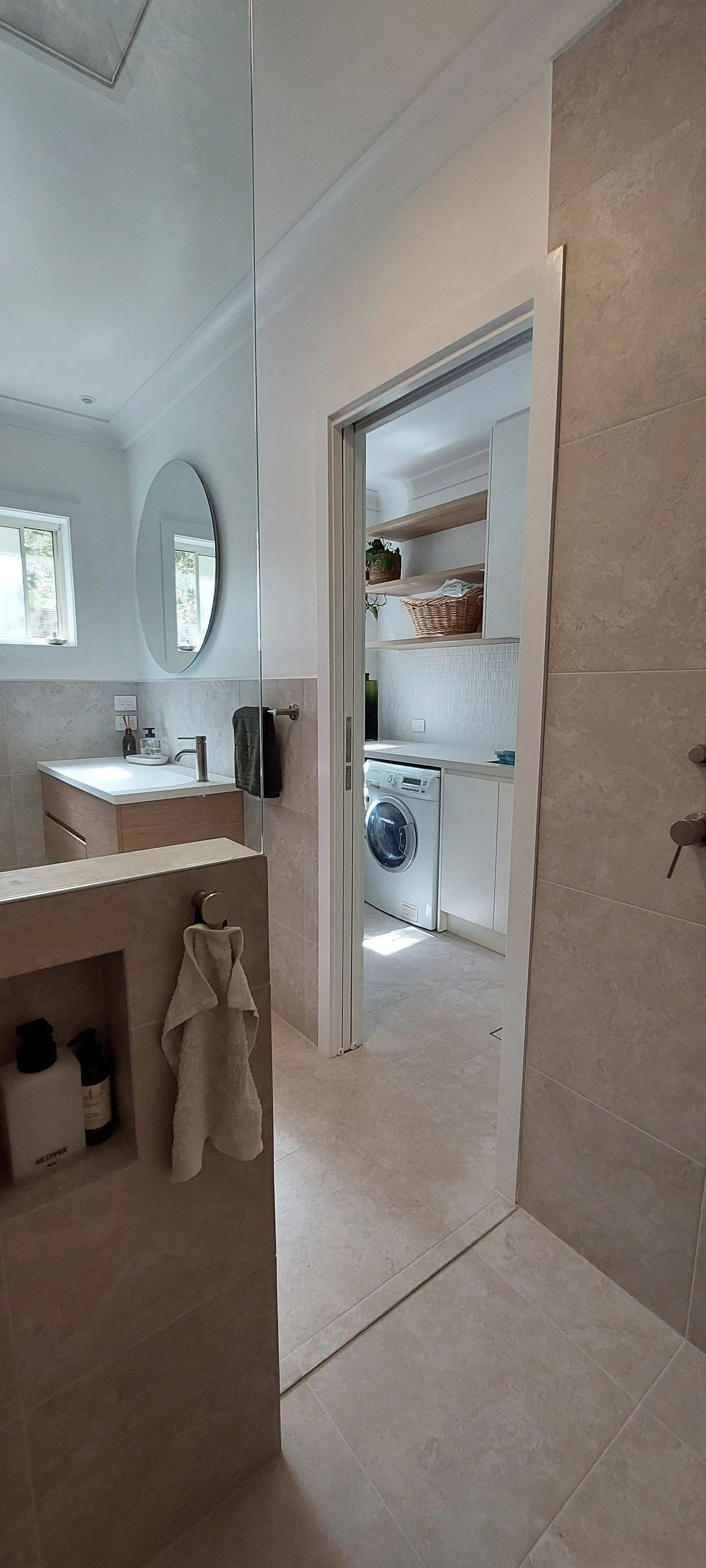 Bathroom and laundry room with a front-loading washing machine, shelves with basket and decor, beige tiled floors, and a mirror reflecting the bathroom sink and window.