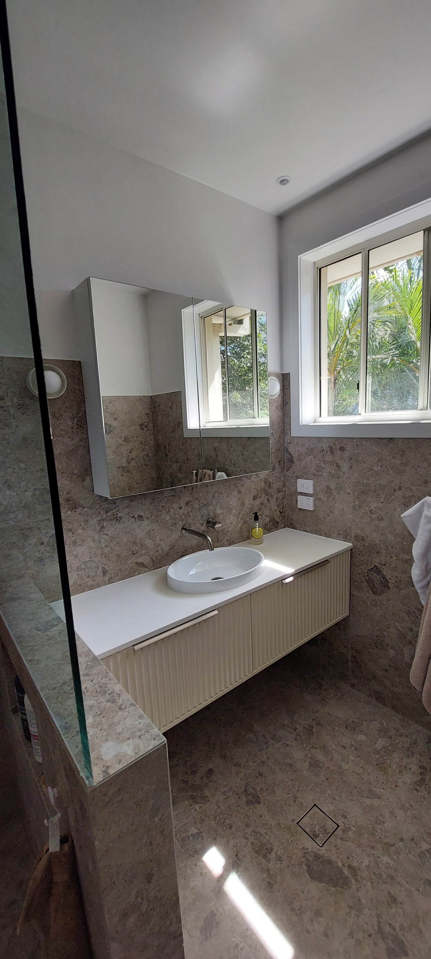 Bathroom with marble walls and floor, a white countertop with an oval sink, a mirror cabinet, and a window showing greenery outside.