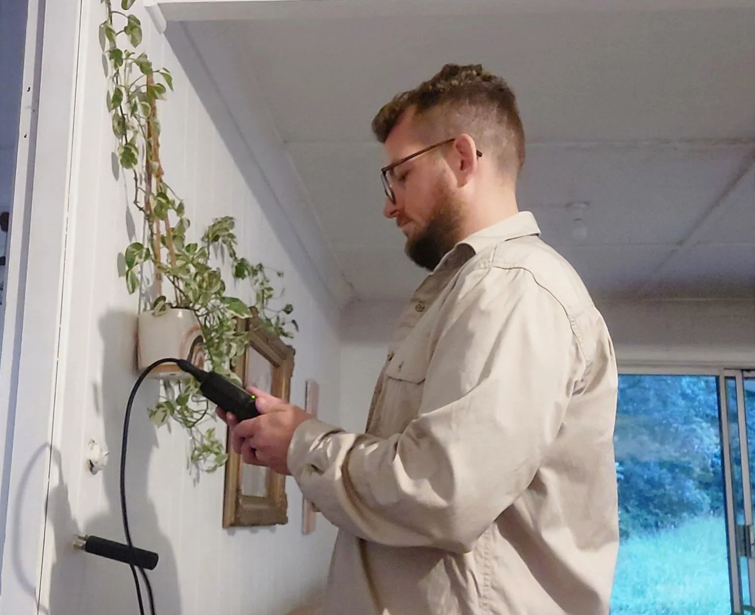 A man wearing glasses and a beige shirt is standing indoors, looking at his phone while connected to an electrical outlet on the wall with a black charger.