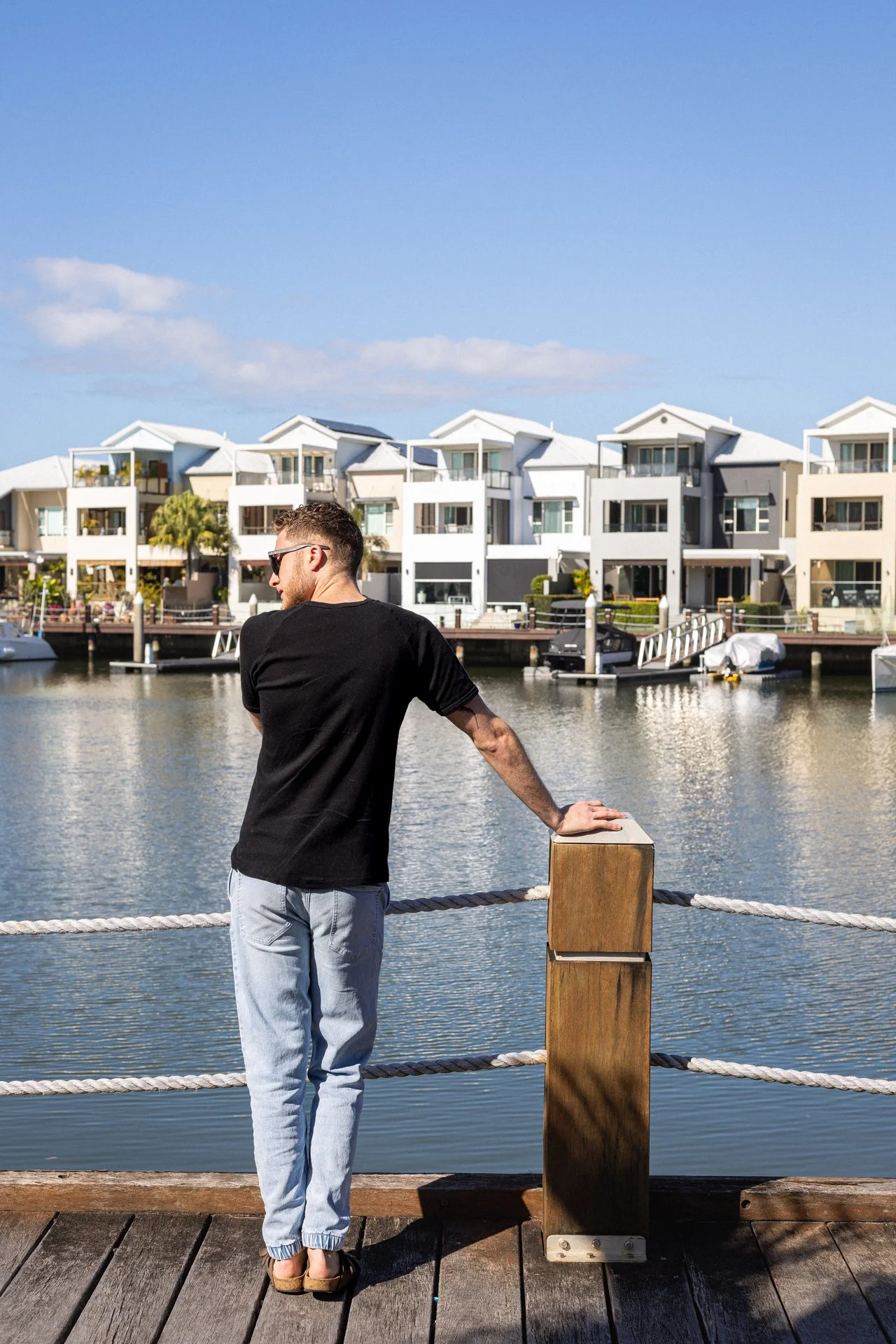 Full-body portrait of man at Coomera Waters boardwalk facing the marina with hands on railing.