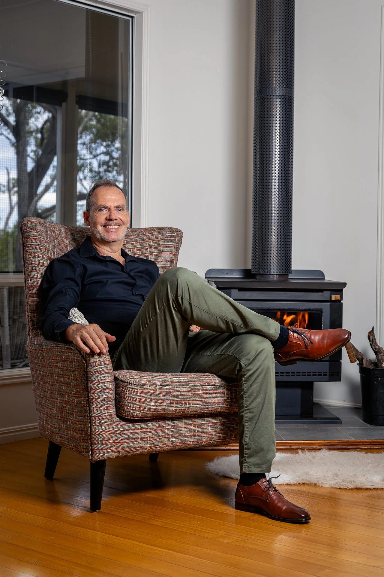 A smiling man with gray hair and a beard, wearing a black shirt and green pants, sitting comfortably in a patterned armchair in front of a fireplace, with a window behind him showing trees outside.