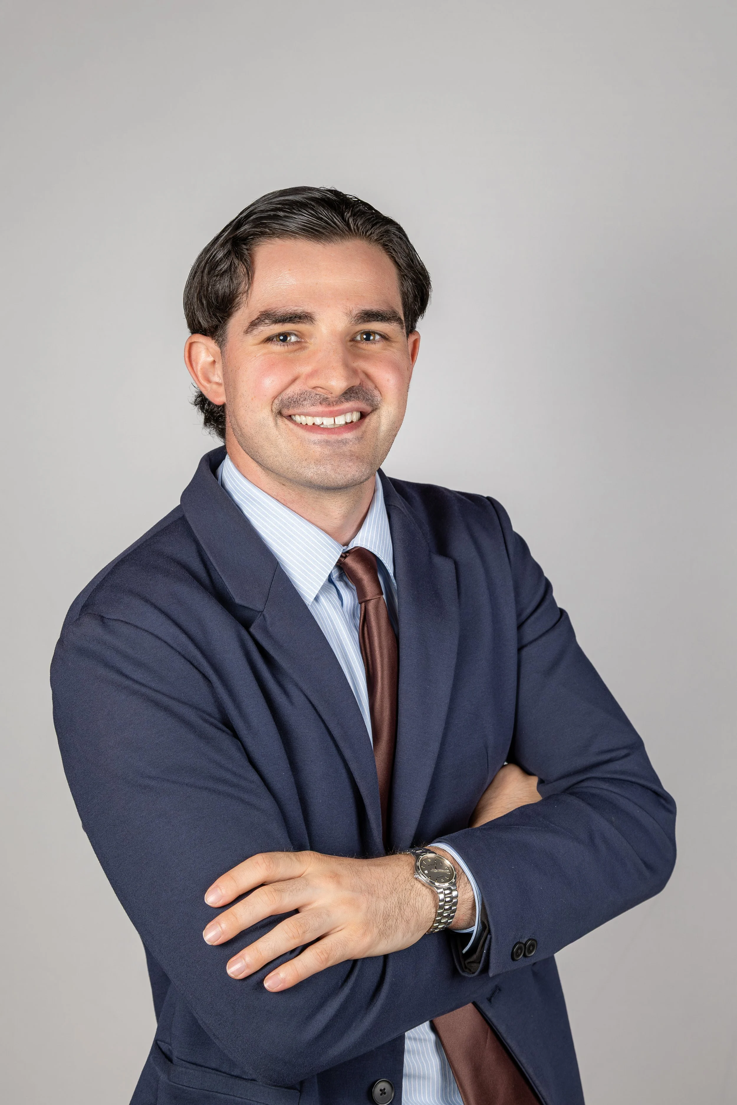 Headshot of a smiling man with dark hair, wearing a navy suit, light blue dress shirt, and a brown tie, posing with arms crossed against a plain light gray background.
