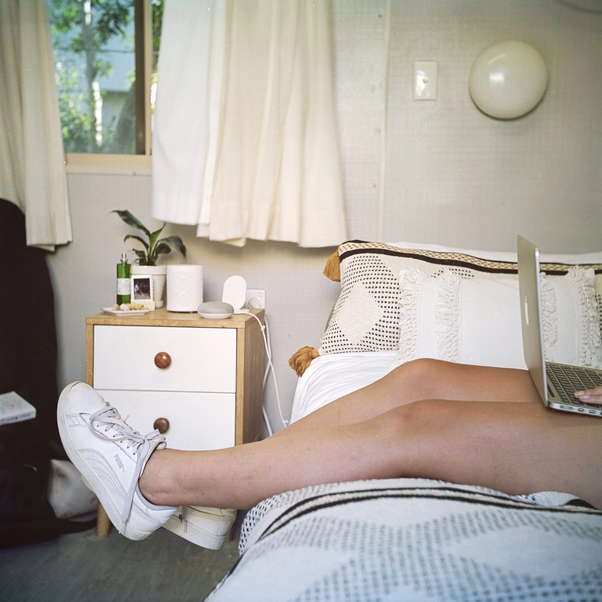 Person lying on a bed using a laptop, wearing white sneakers. There's a bedside table with a plant and small electronic devices next to a window with curtains.