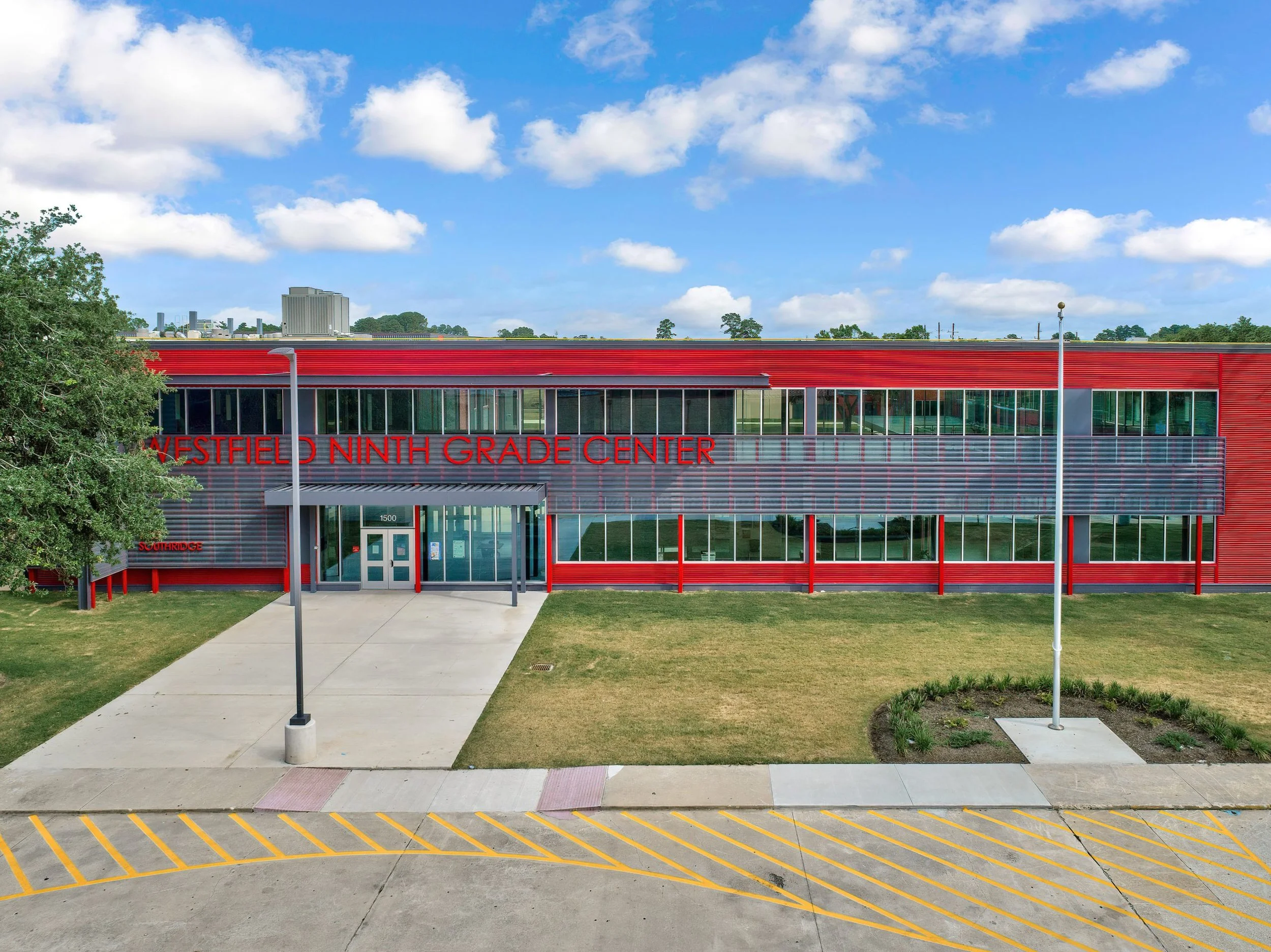 Exterior view of Westfield Ninth Grade Center, a modern red school building with large glass windows and a main entrance under a small canopy, surrounded by a grass lawn, flagpoles, and a parking lot with yellow stripes, under a partly cloudy sky.