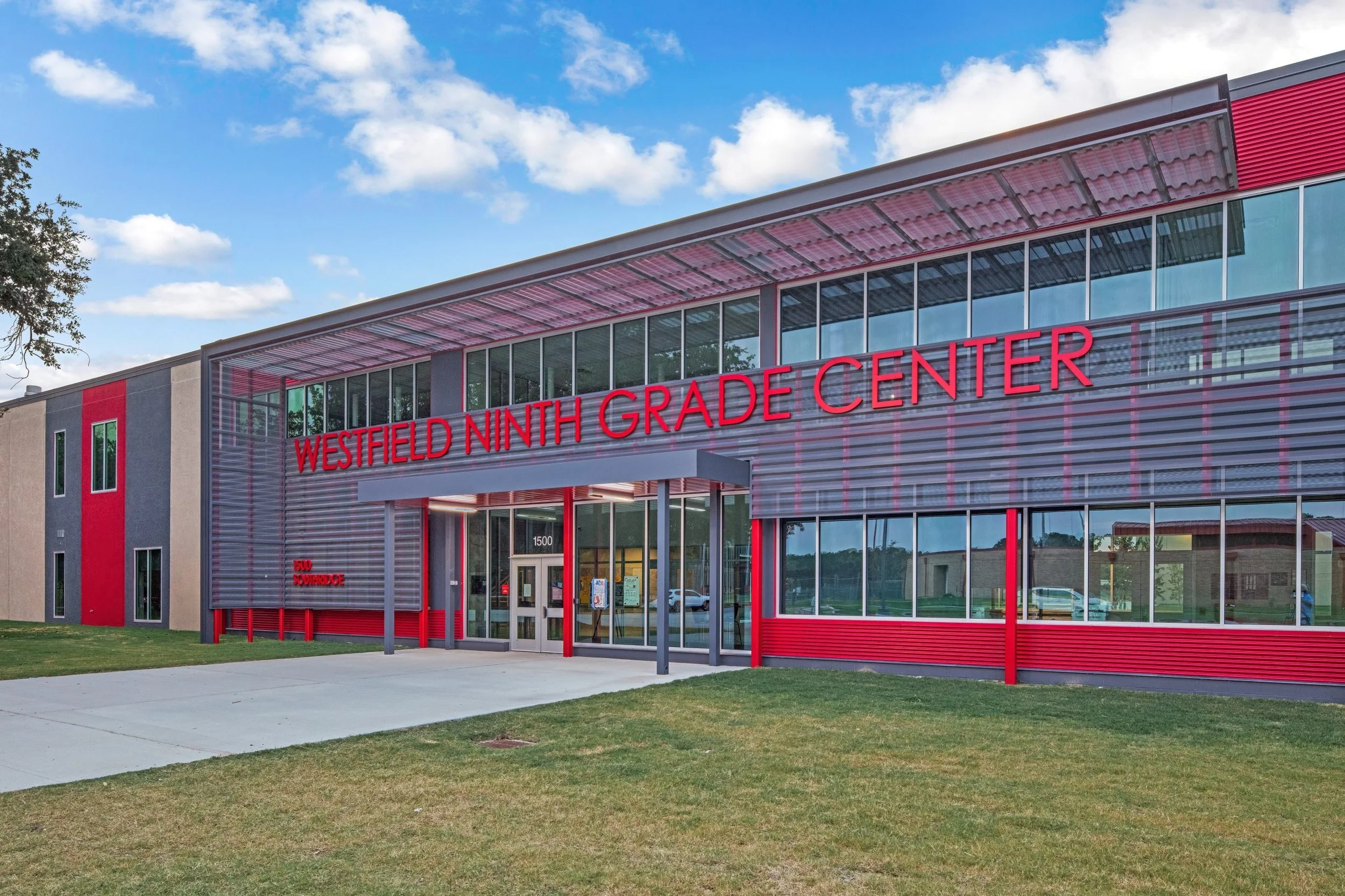 Exterior view of Westfield Ninth Grade Center school building with a red and gray facade, large windows, and a sign displaying the school's name in red letters. The sky is partly cloudy and there is a grassy area in front of the building.
