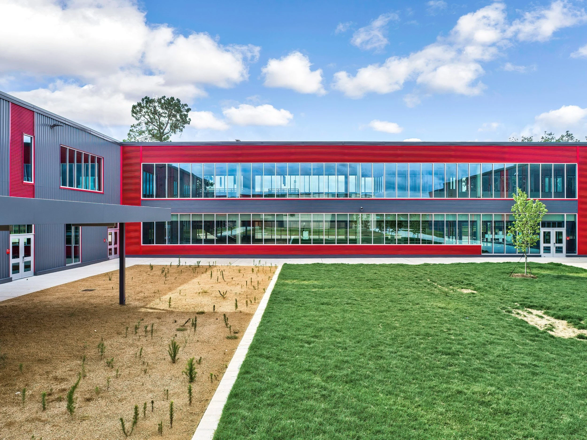 Modern school building with red and gray exterior, large glass windows, a small planted area, a grassy field, and a young tree in the foreground under a partly cloudy sky.