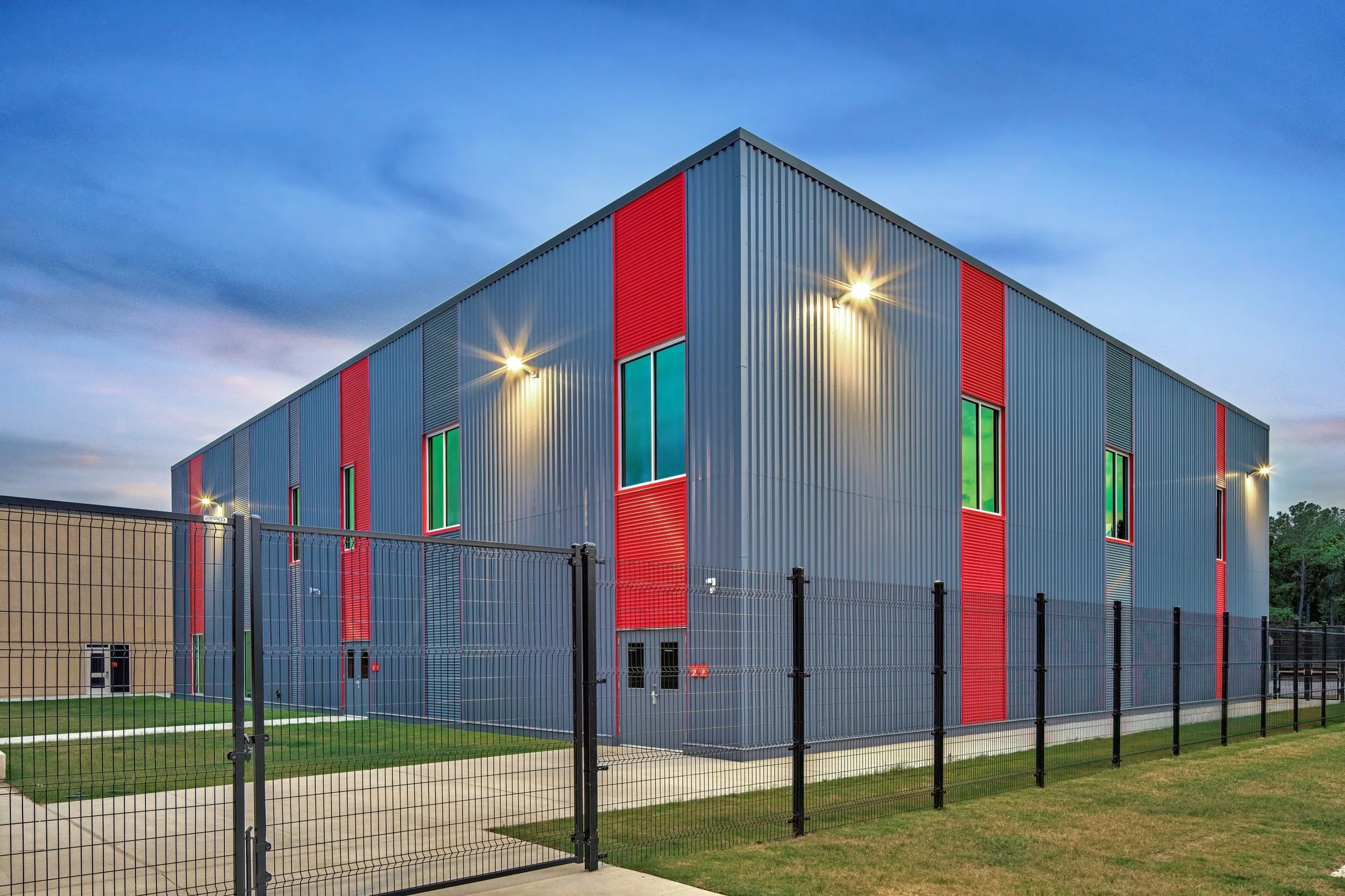 Modern gray and red commercial or industrial building with windows, surrounded by a black metal fence and lit by exterior lights, during evening with cloudy sky.