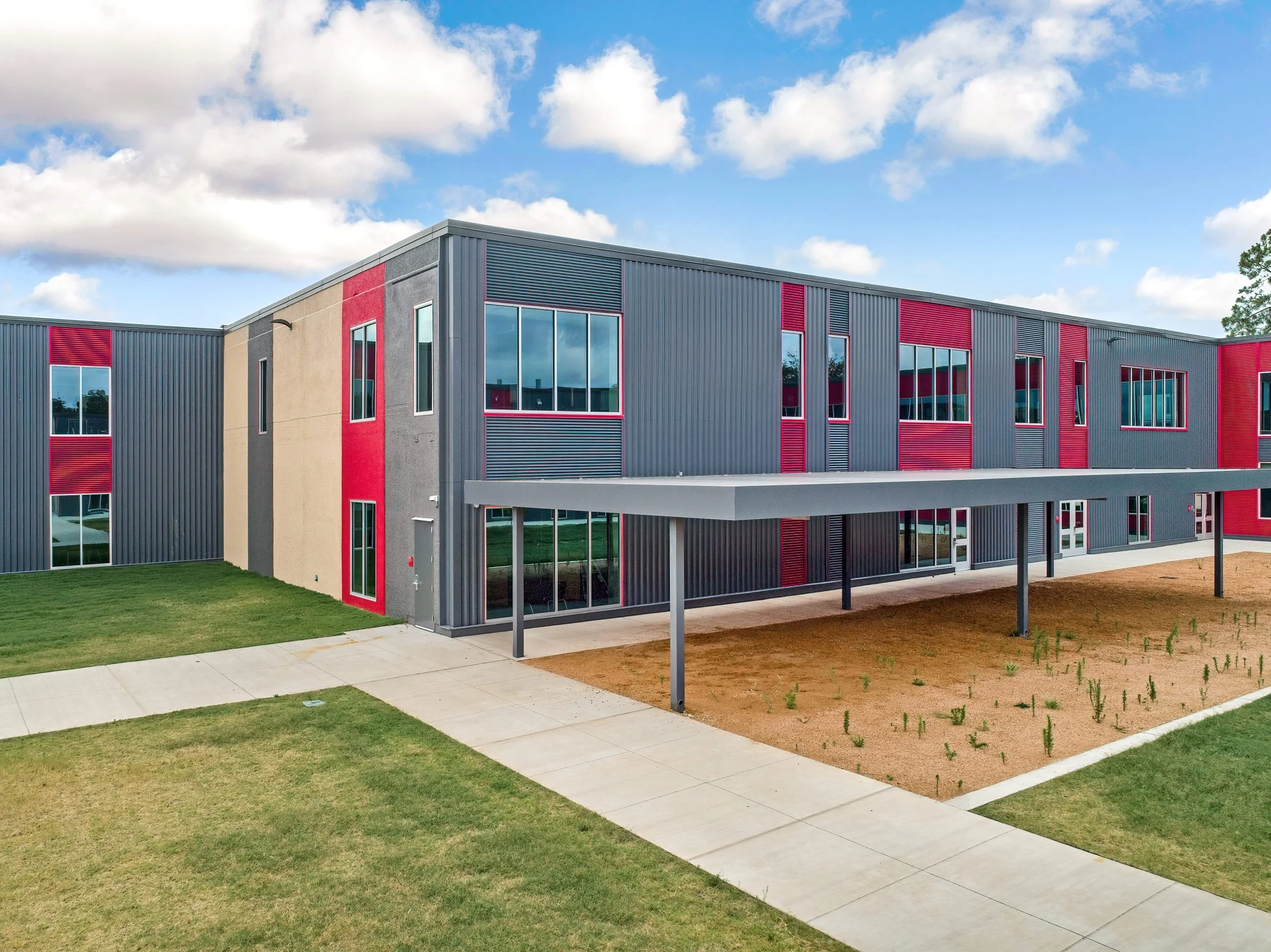 Modern two-story school building with gray and red metal siding, green lawn, concrete walkway, and a covered entrance, under a partly cloudy sky.