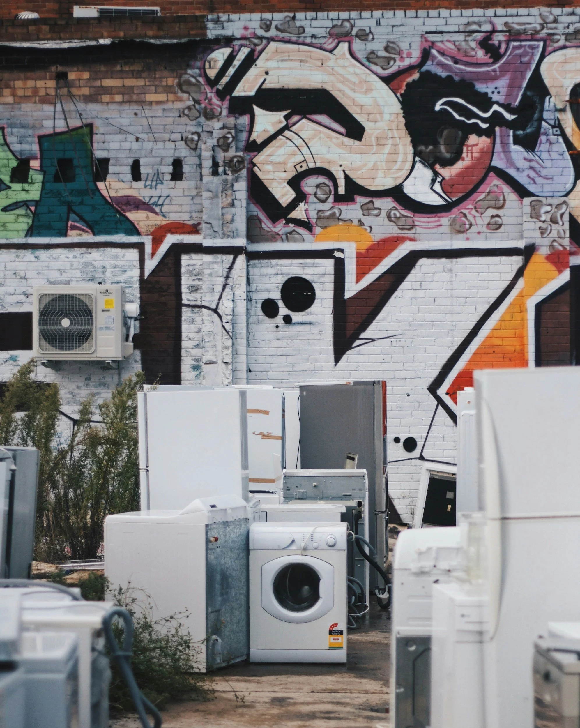 A graffiti mural on a brick wall featuring a stylized human face wearing sunglasses, surrounded by abstract shapes and designs, with various washing machines and appliances placed in front of the wall.