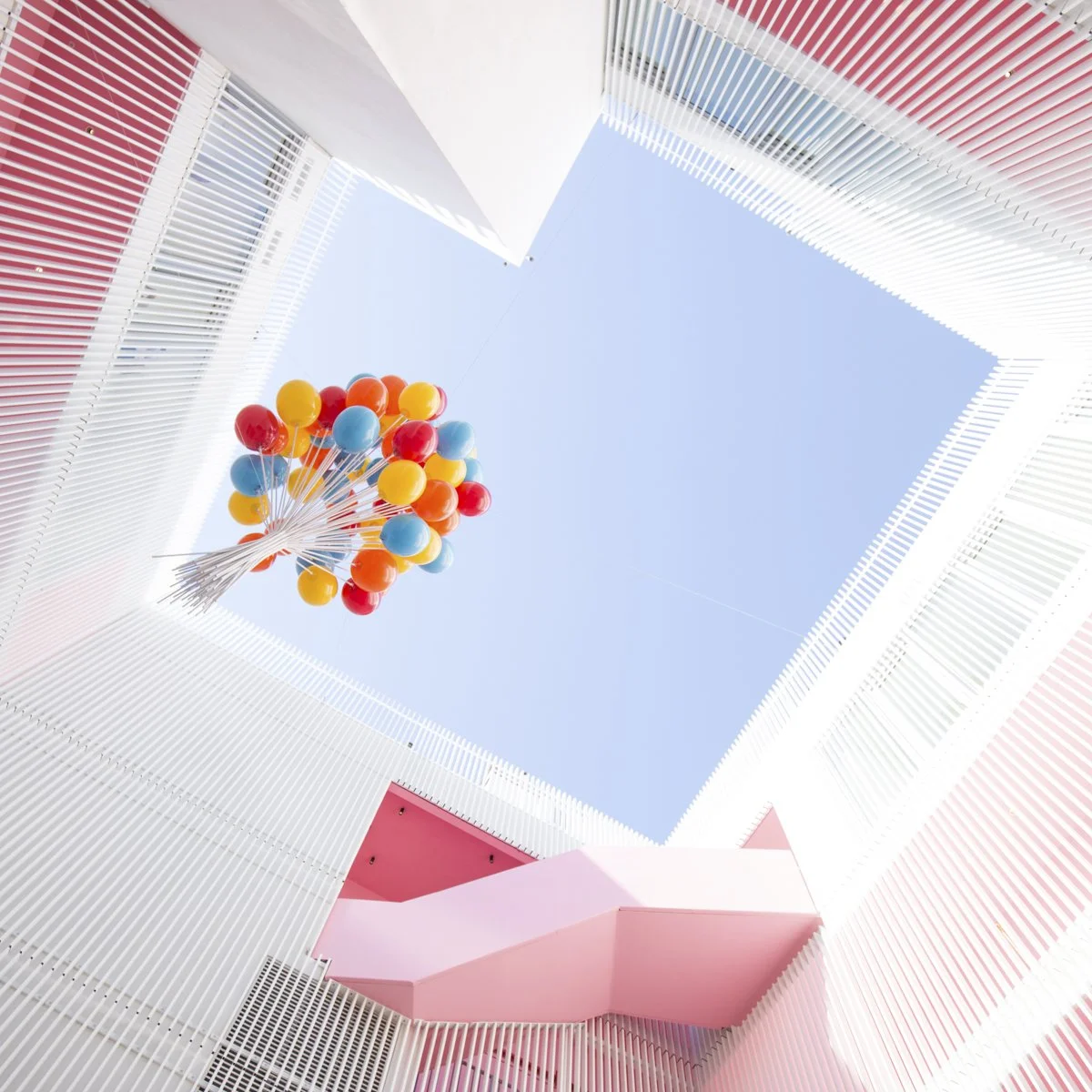 Looking up at a colorful balloon arrangement hanging in an atrium with a clear blue sky visible through the open ceiling, surrounded by white and pink modern architectural walls.