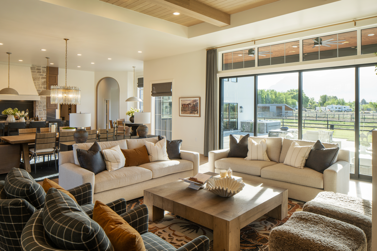 Living room with beige sofas, plaid armchairs, a wooden coffee table, large windows with city view, and an open dining area in the background.