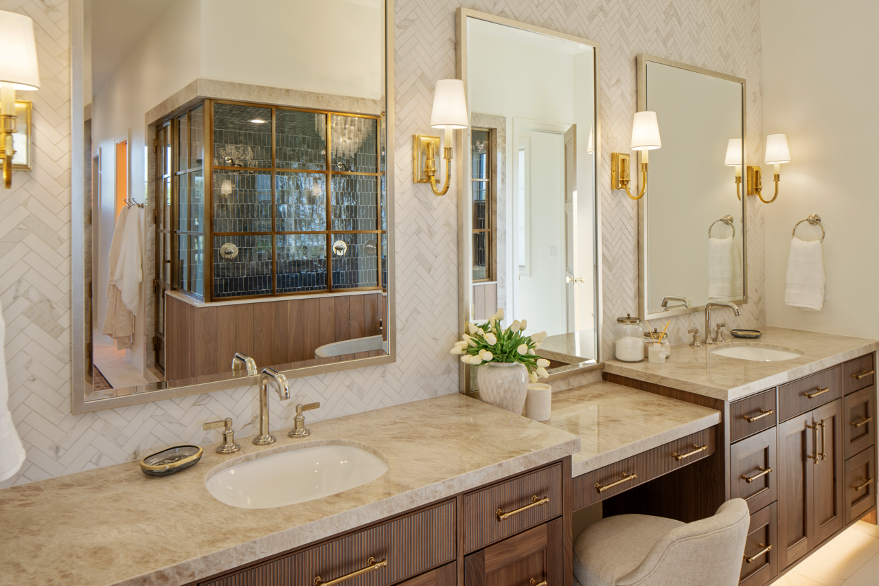 Bathroom vanity with two sinks, large mirrors, brass fixtures, and wall-mounted lights, with a decorative flower arrangement on the countertop.