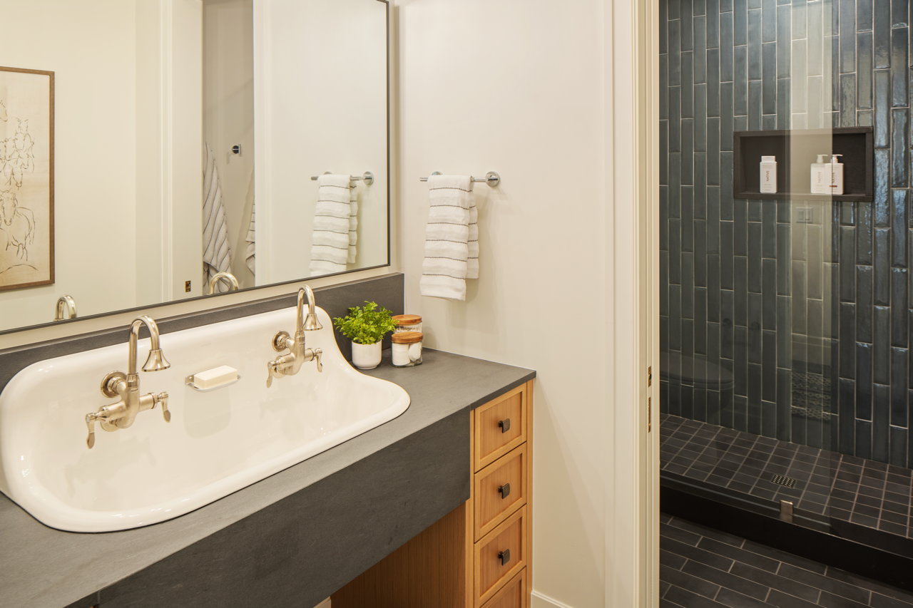 Bathroom with a white sink and a large mirror, with a shower area visible in the background, featuring dark gray tiles and a built-in shelf.