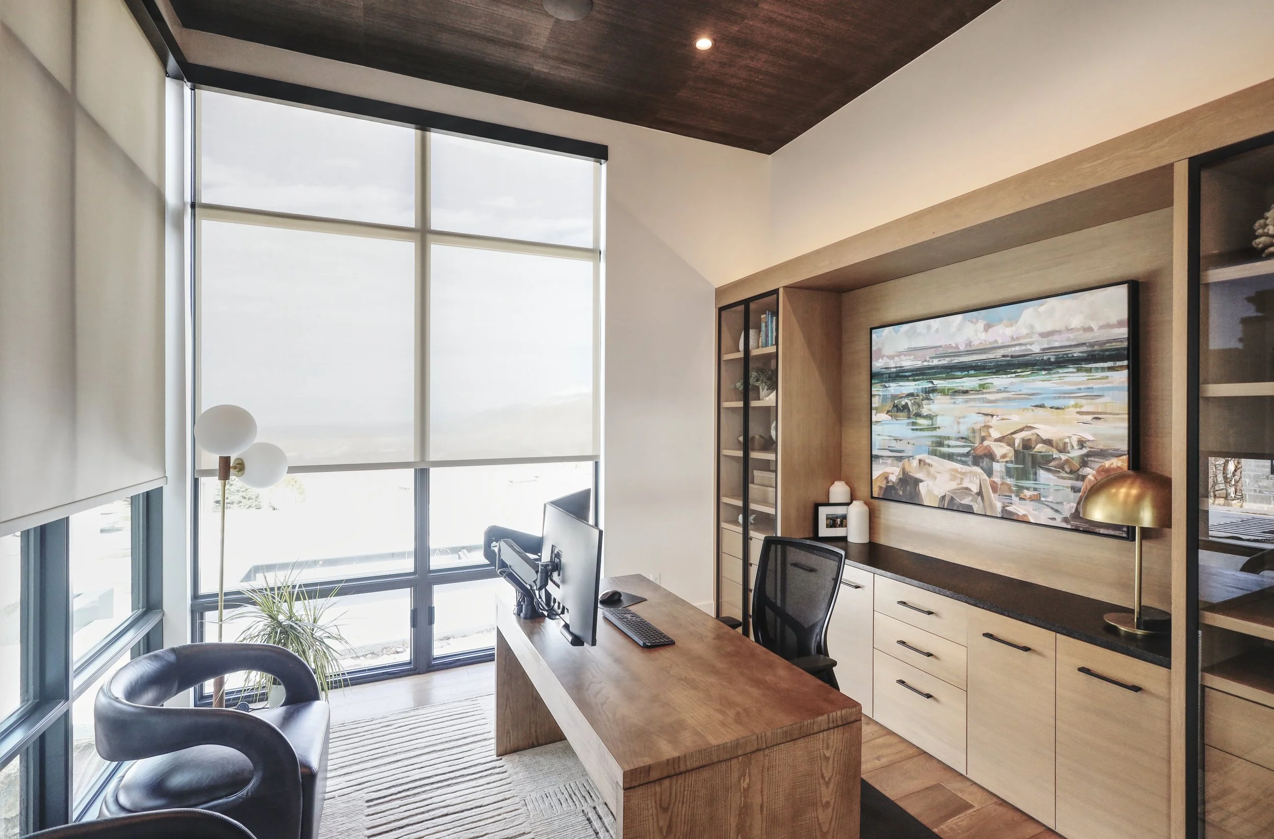 Modern home office with large window, wooden desk, black office chair, and built-in wooden bookshelf with a large landscape painting above it.