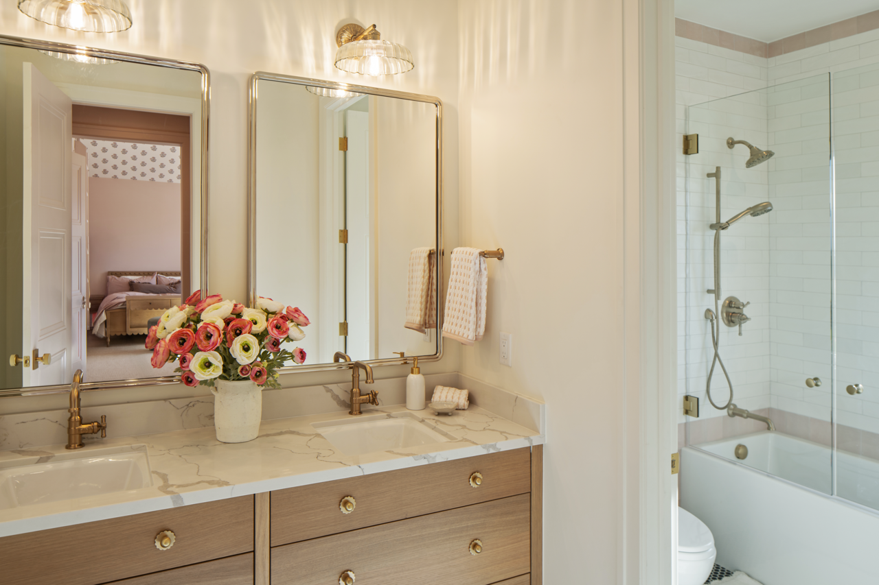 Bathroom with a double vanity featuring a marble countertop, a vase of pink and white flowers, and large mirrors. There's a towel rack with two towels and a shower/tub combination with a glass door.