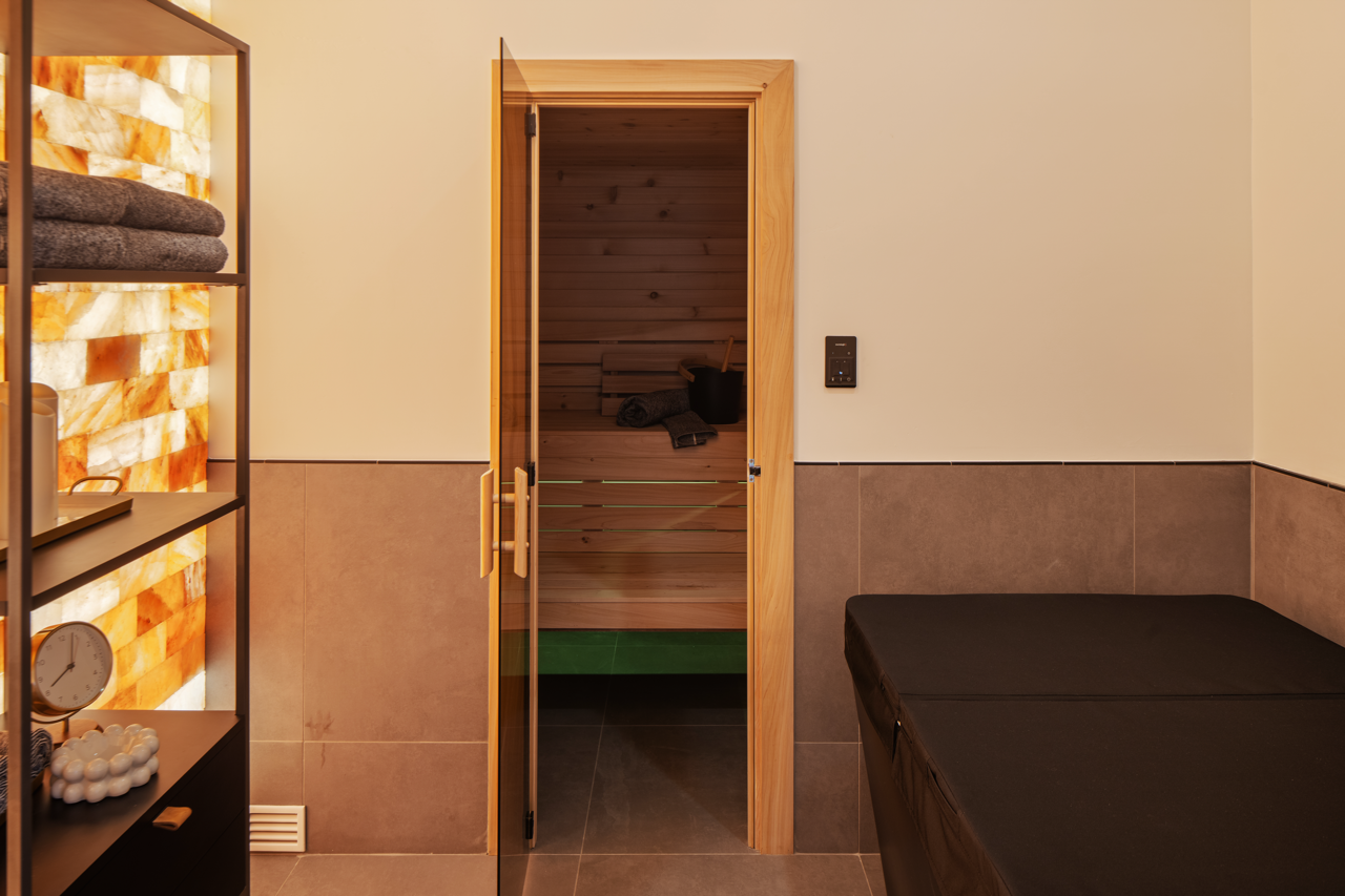 A room with a door leading to a sauna, featuring a wooden interior, a black bucket, and rolled towels. The room has beige walls with a darker tiled lower half, a black massage table, and a black shelf with towels, a clock, and bath salts.