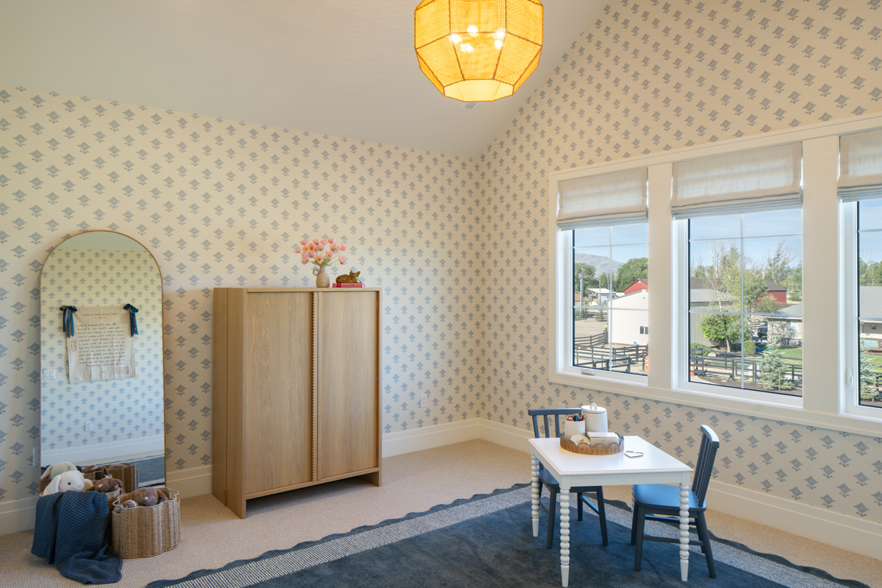 Child's playroom with wallpaper, window, small table and chairs, wardrobe, basket of stuffed animals, and a mirror.