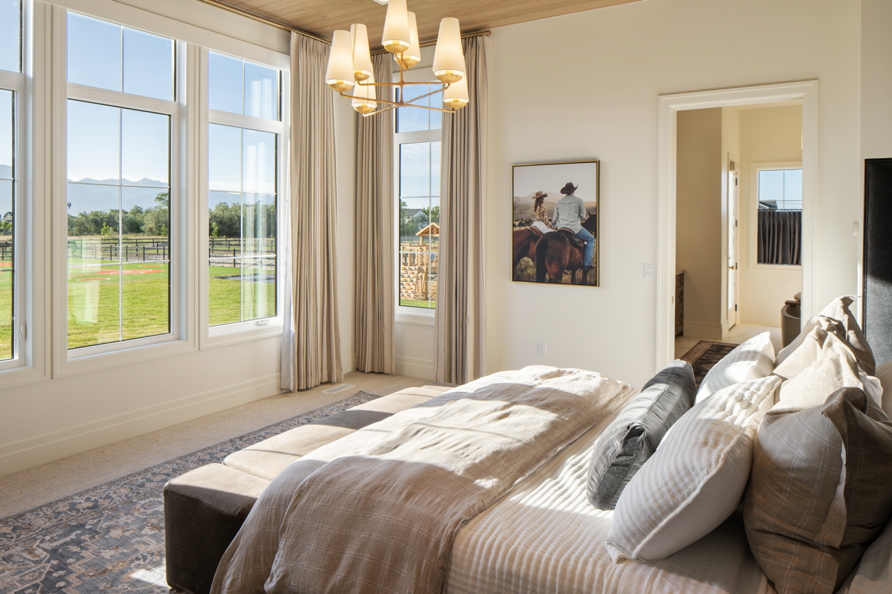 Bright bedroom with large windows overlooking green fields and a distant mountain range, decorated with neutral-toned bedding, a chandelier, and a framed picture on the wall.