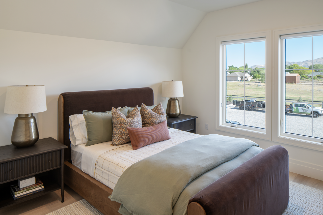 A bedroom with a brown upholstered bed, light green and floral pillows, white bedding, two black nightstands with matching lamps, and a large window showing a view of a yard, parked trucks, and distant mountains.