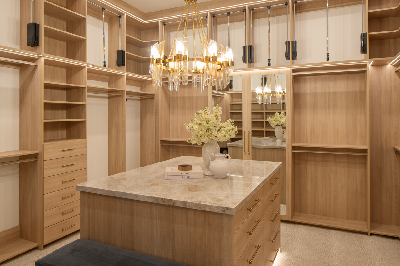 Beautiful walk-in closet with wooden shelves, drawers, and hanging space, a large marble-topped island center, floral decor, and a gold chandelier light fixture.
