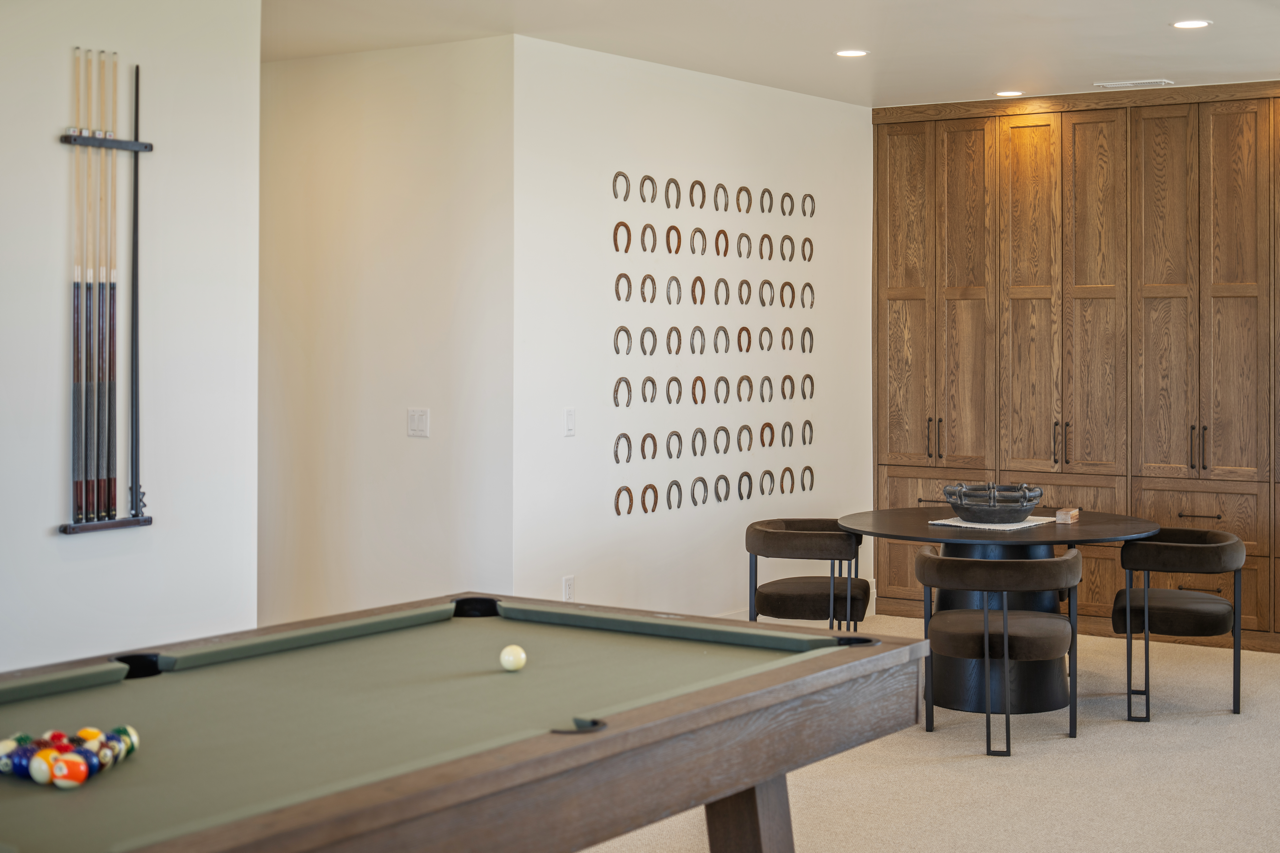 Room with a pool table, wall-mounted cue rack, decorative horseshoe display, round table with four chairs, and wooden built-in cabinets.