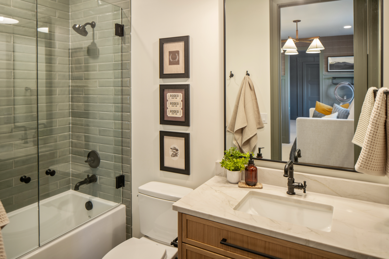 Bathroom with a walk-in shower, a toilet, a wooden vanity with a marble countertop, a mirror, and wall art. The shower has green tiles and black fixtures, and the vanity has a plant, soap dispenser, and hand towel.