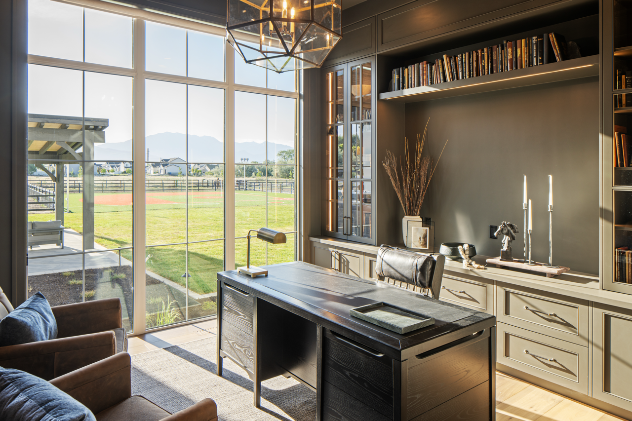 A modern home office with large windows overlooking a grassy outdoor area and tennis court. The office has a black desk, a leather chair, built-in bookshelves, and decorative items including candles and plants.