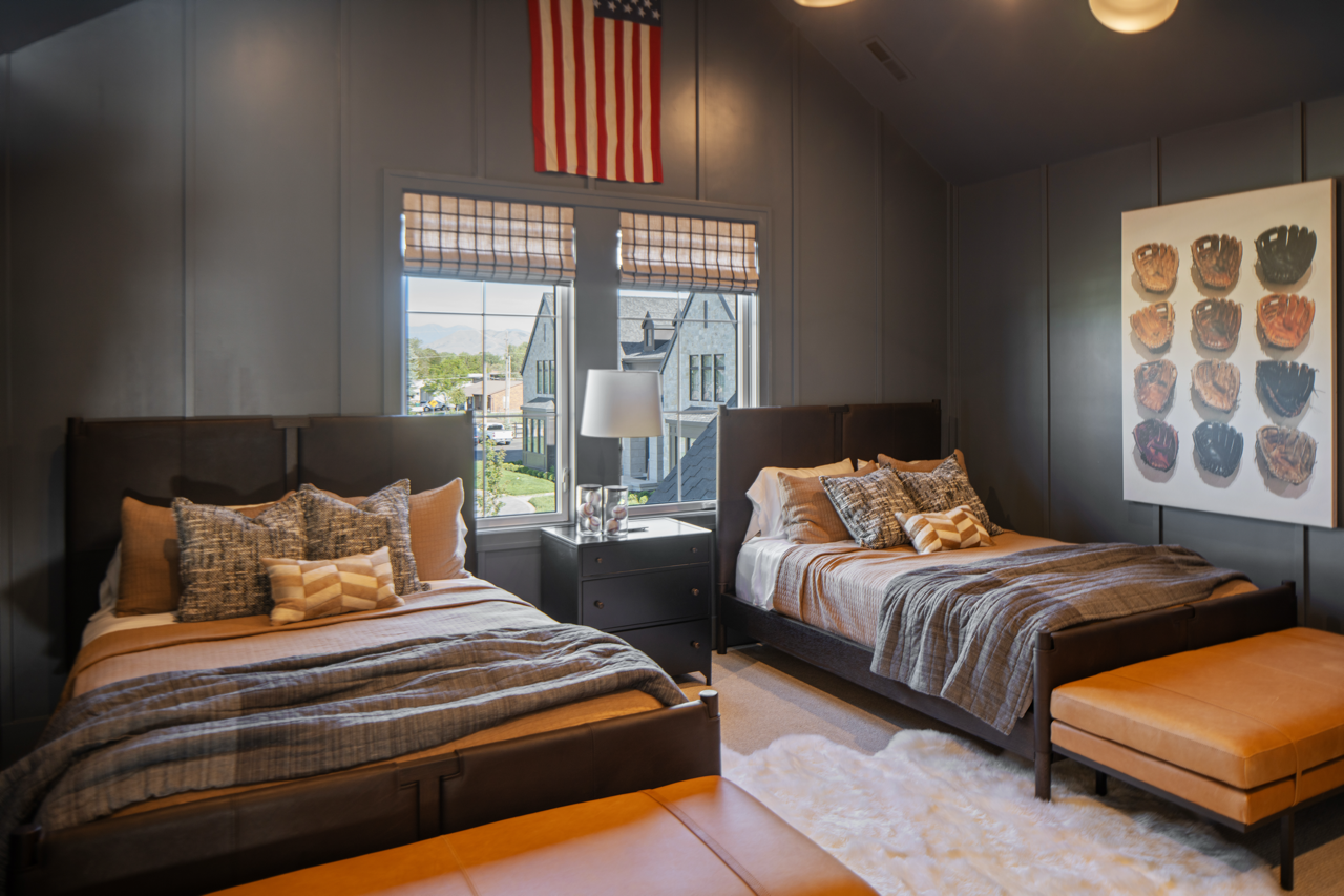 A bedroom with two beds, a nightstand with a lamp, and a window showing houses outside. There is wall art of baseball gloves above the right bed and an American flag on the wall.