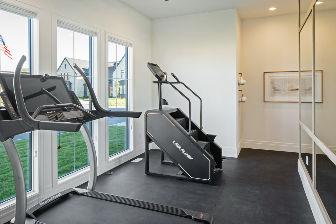 Home gym with a treadmill and an elliptical machine near large windows showing a yard and houses outside.