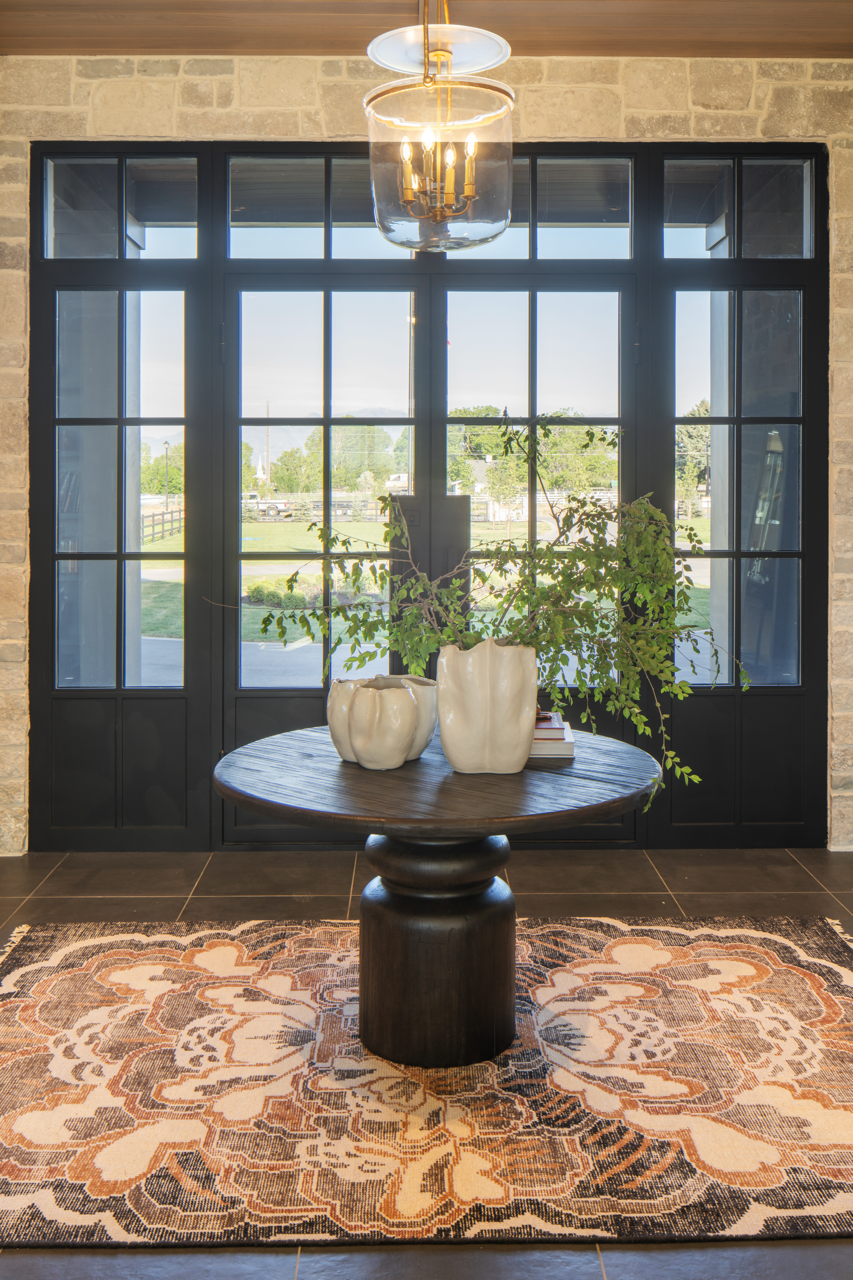 A round wooden table with three white ceramic vases containing greenery, set on an ornate patterned rug. Behind the table are large black-framed glass doors showing an outdoor scene with trees and a clear sky. Above, a modern chandelier with exposed 