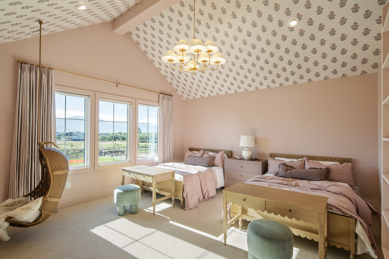 Bright bedroom with vaulted ceiling, floral wallpaper on ceiling, two beds with pink bedding, a white bedside table with a lamp, a hanging wicker chair, a small green ottoman, and a large window showing a ranch landscape.
