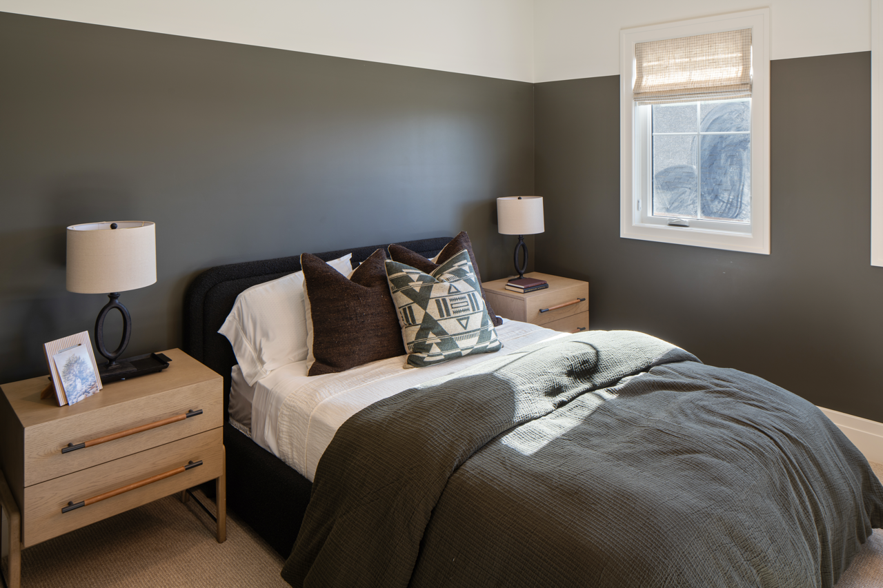 A bedroom featuring a bed with white sheets, a dark bed frame, and multiple pillows. Two bedside tables with lamps flank the bed. A window with a beige curtain is on the right, allowing sunlight to softly illuminate the room.