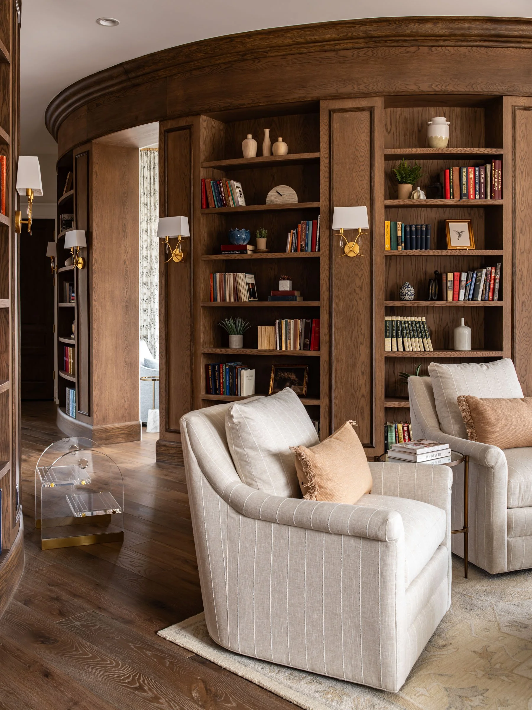Living room with beige striped armchair, pillows, wooden bookshelf, hardwood flooring, and wall-mounted lamps.