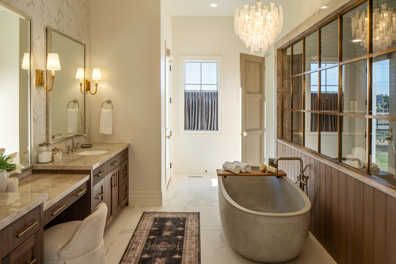 A luxurious bathroom featuring a large bathtub with a wooden tray holding towels and bath accessories, a marble countertop vanity with two mirrors and brass wall sconces, a window with striped curtains, a patterned rug, and a wooden framed glass wall