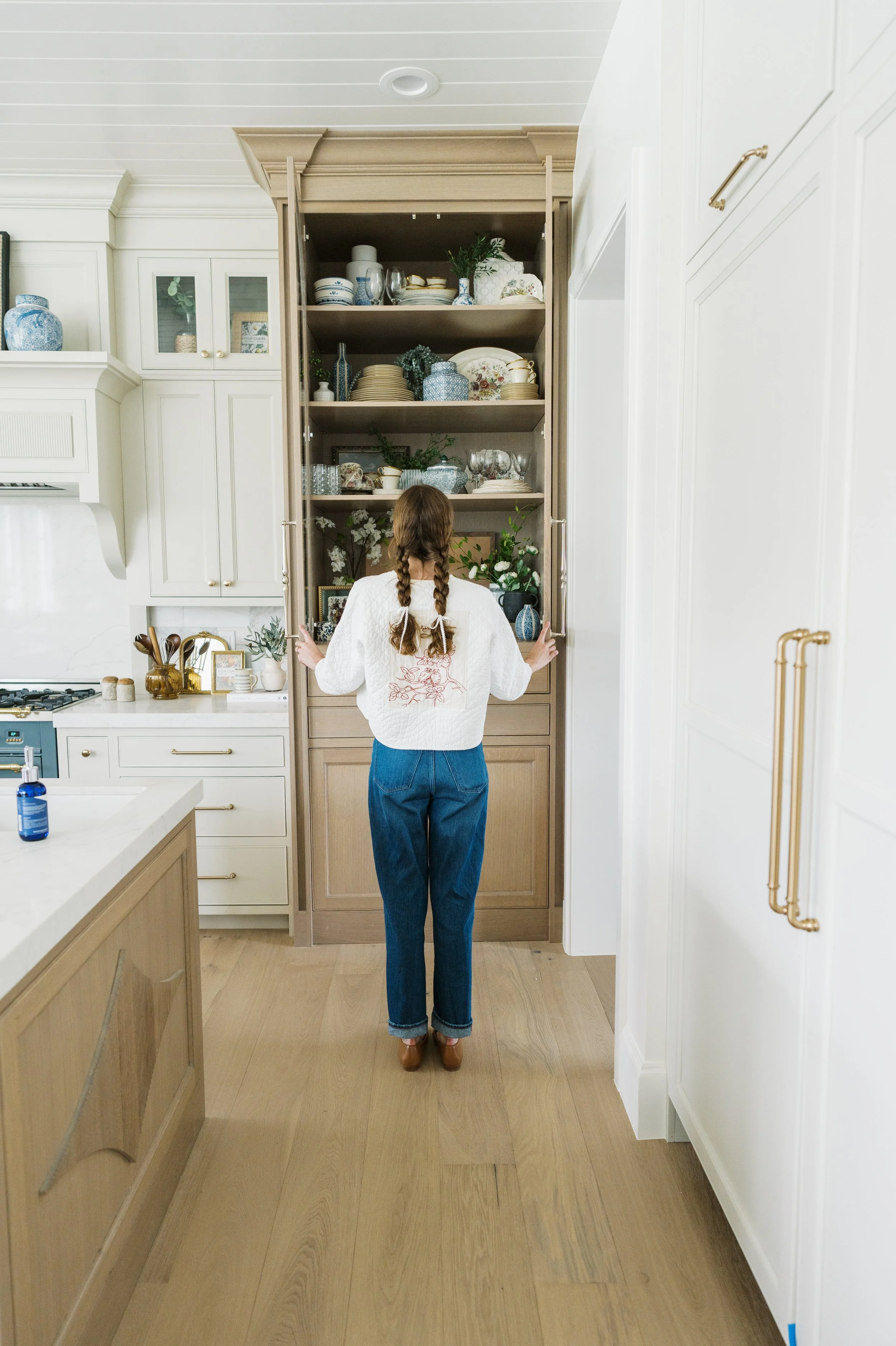 A person with braided hair and jeans standing in front of a large open cabinet filled with dishes, bowls, and decorative items in a bright kitchen.