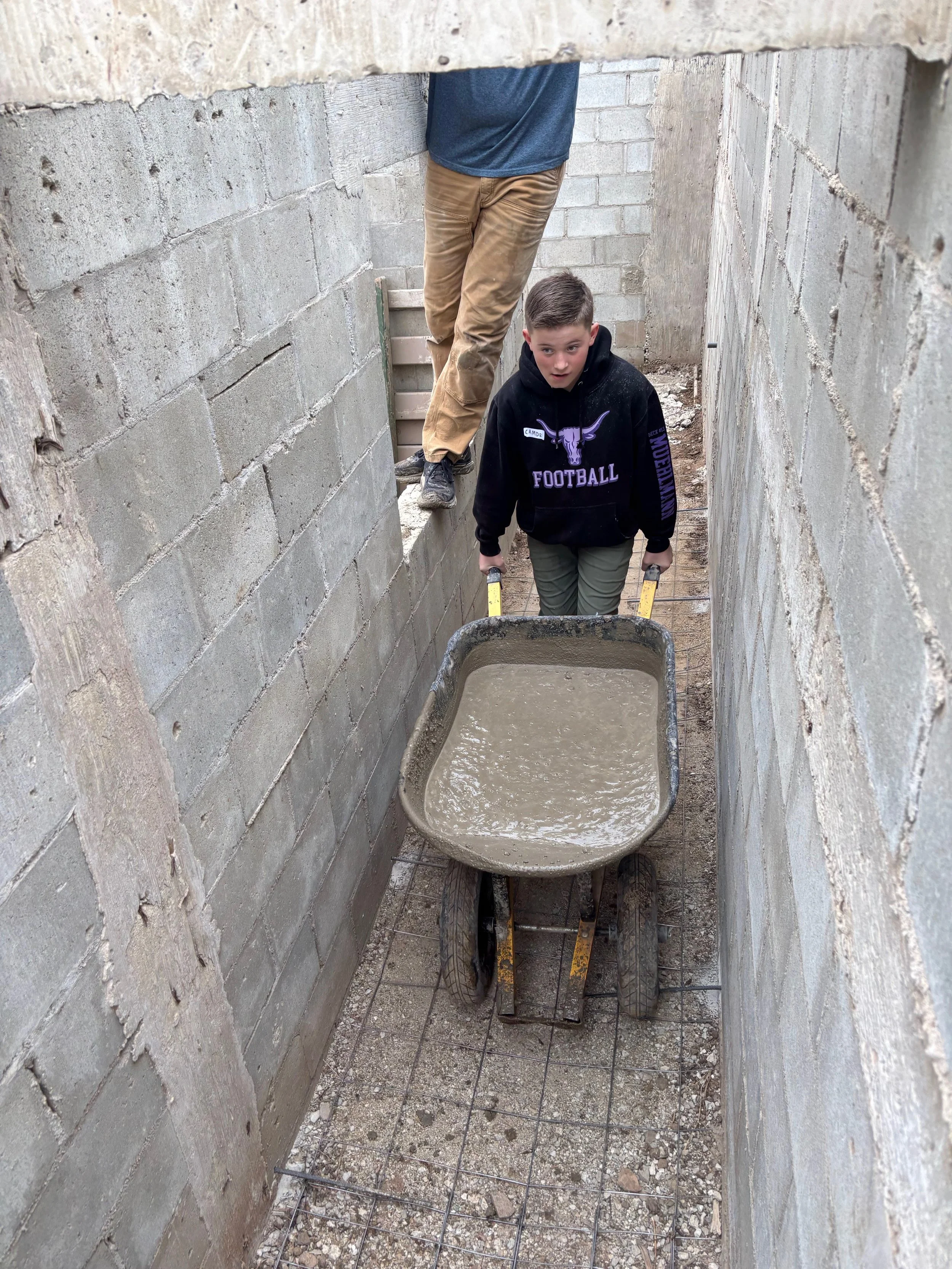 A small construction site with a concrete cinder block wall, a wheelbarrow filled with wet cement, and a person working on the floor, visible from the waist down.